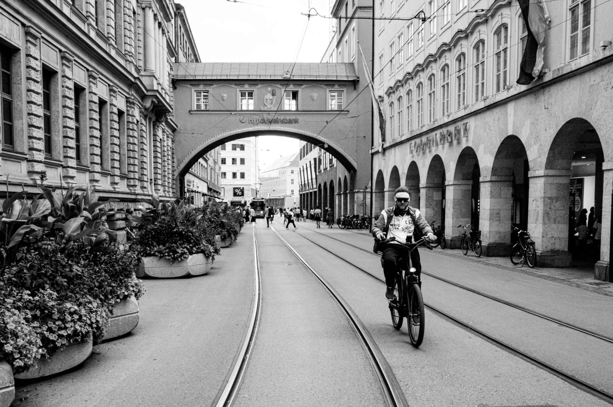Cyclist rides along a historic European street with rail tracks and arches.
