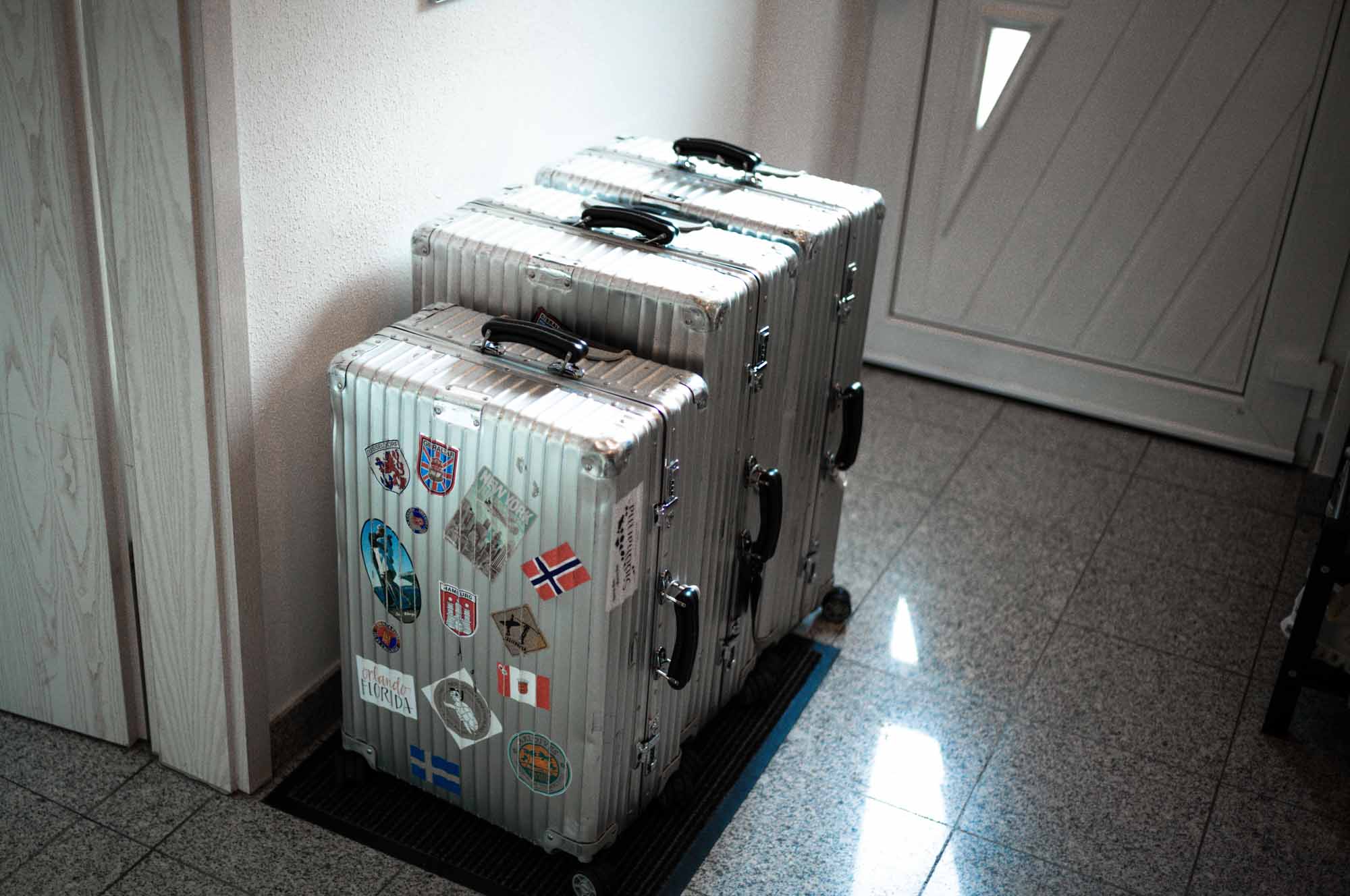 Three silver suitcases with travel stickers lined up against a wall in a well-lit hallway.