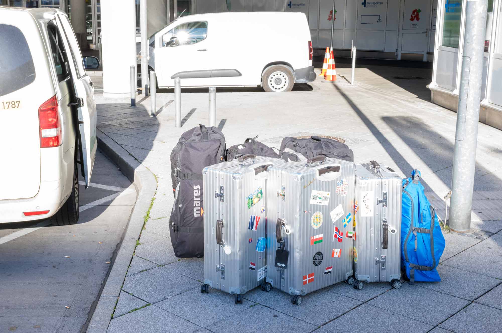 Suitcases and bags with travel stickers on sidewalk, next to parked cars in urban setting.