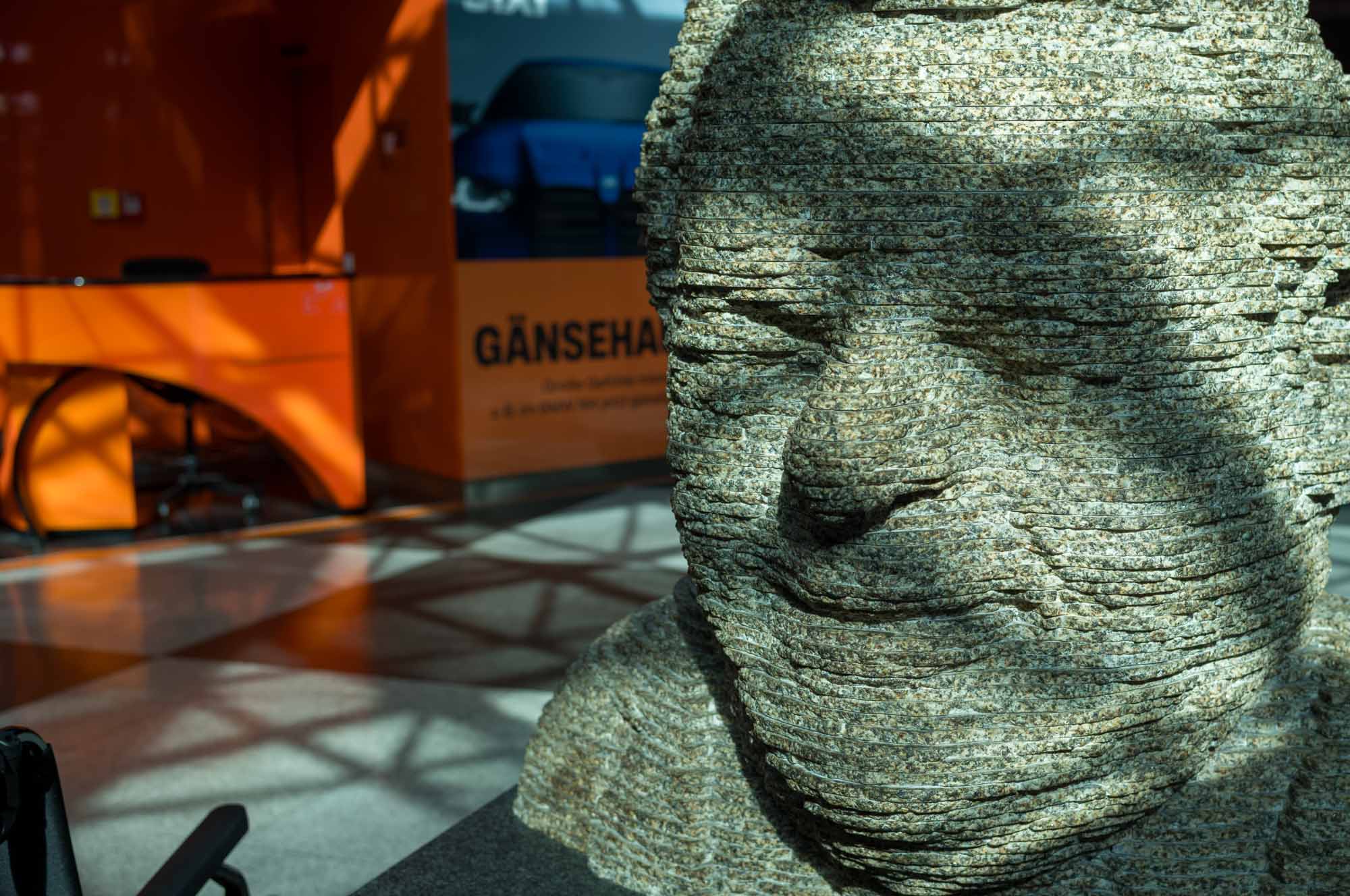 Stone sculpture of a face with textured layers in a sunlit modern interior with orange and blue background elements.