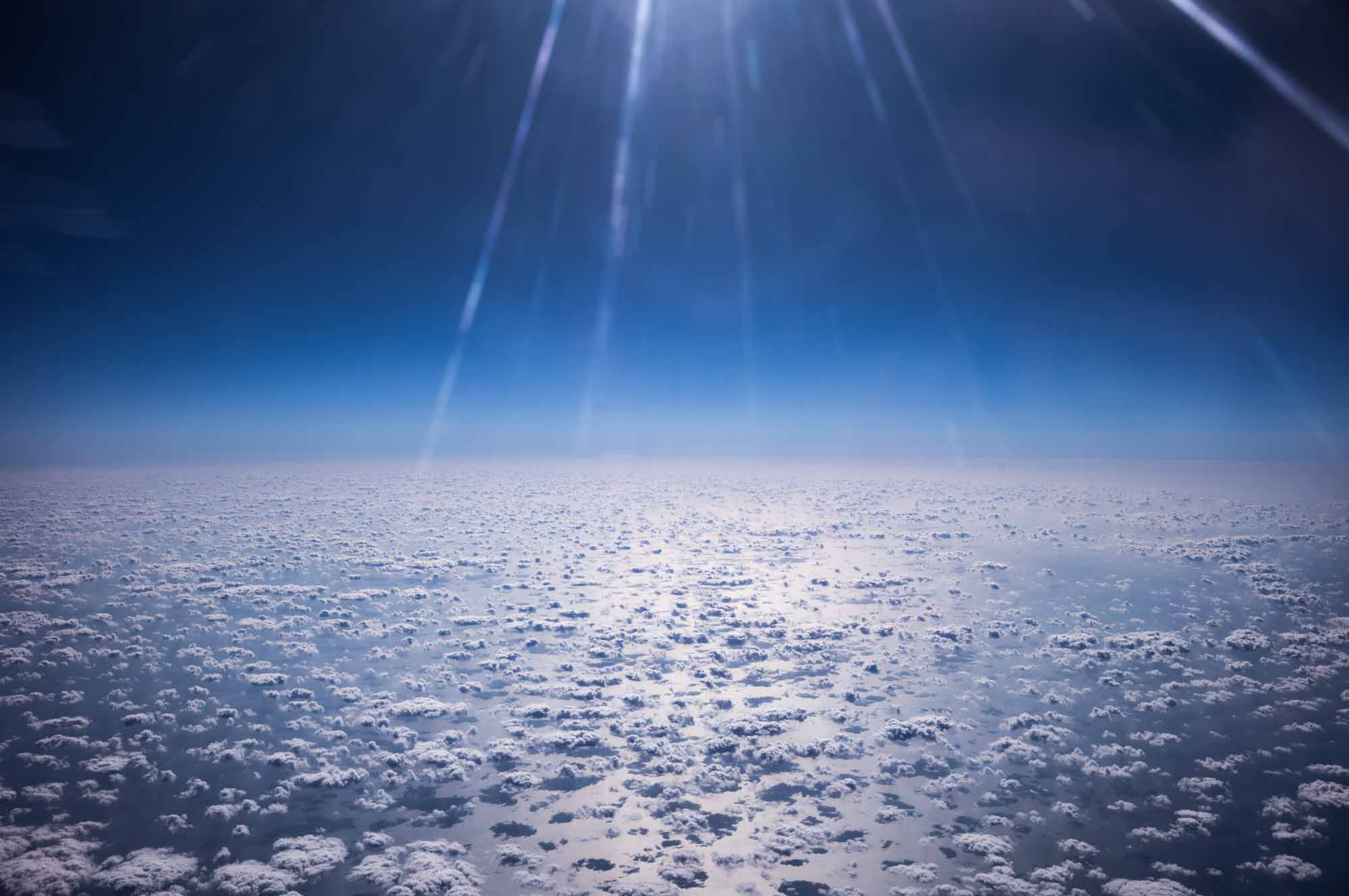 Aerial view of sunlit cloudscape and blue sky with radiant solar rays above, creating a serene and expansive atmosphere.