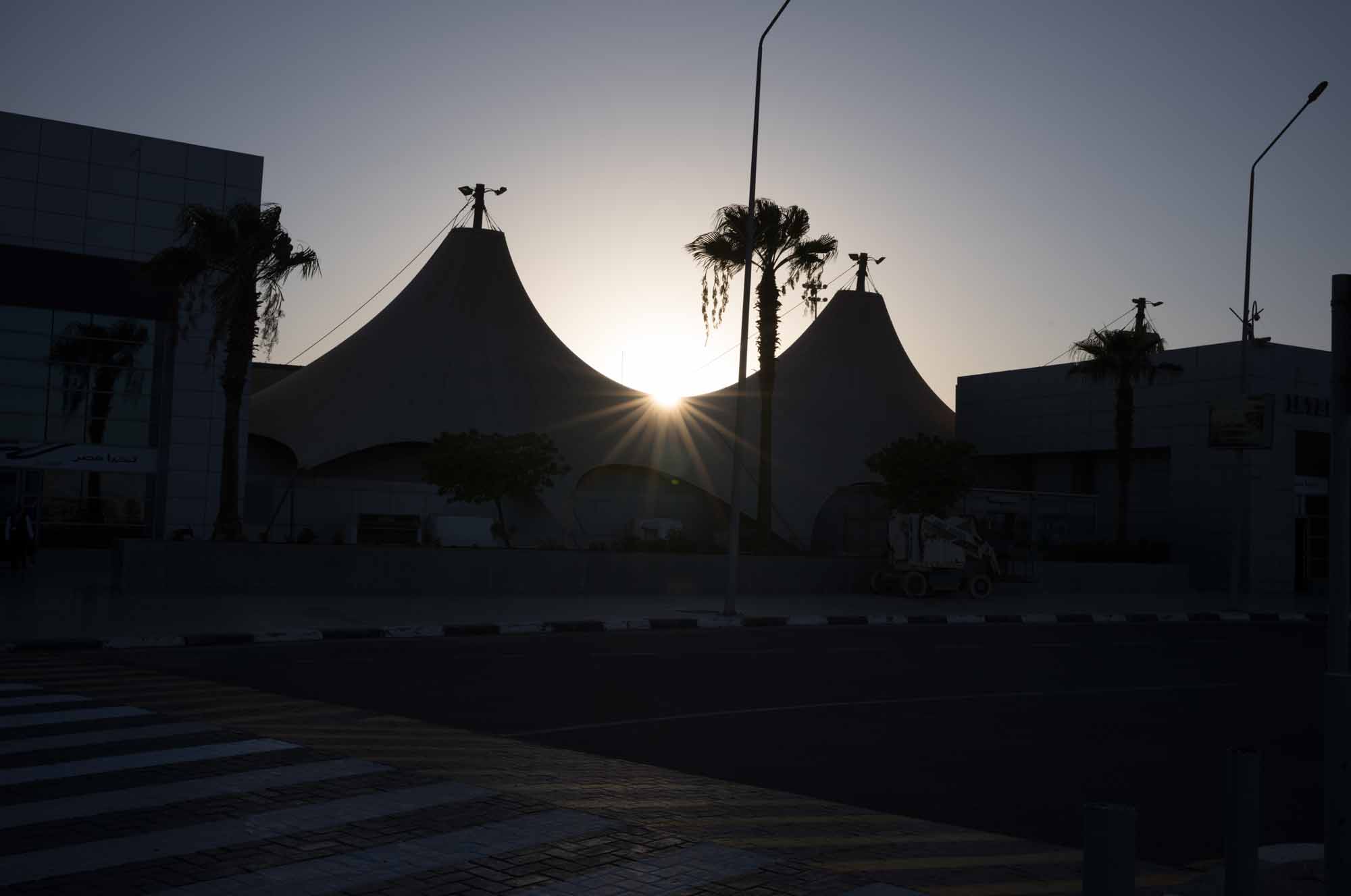 Sunset behind modern architectural tent structures with silhouettes of palm trees and street lights.