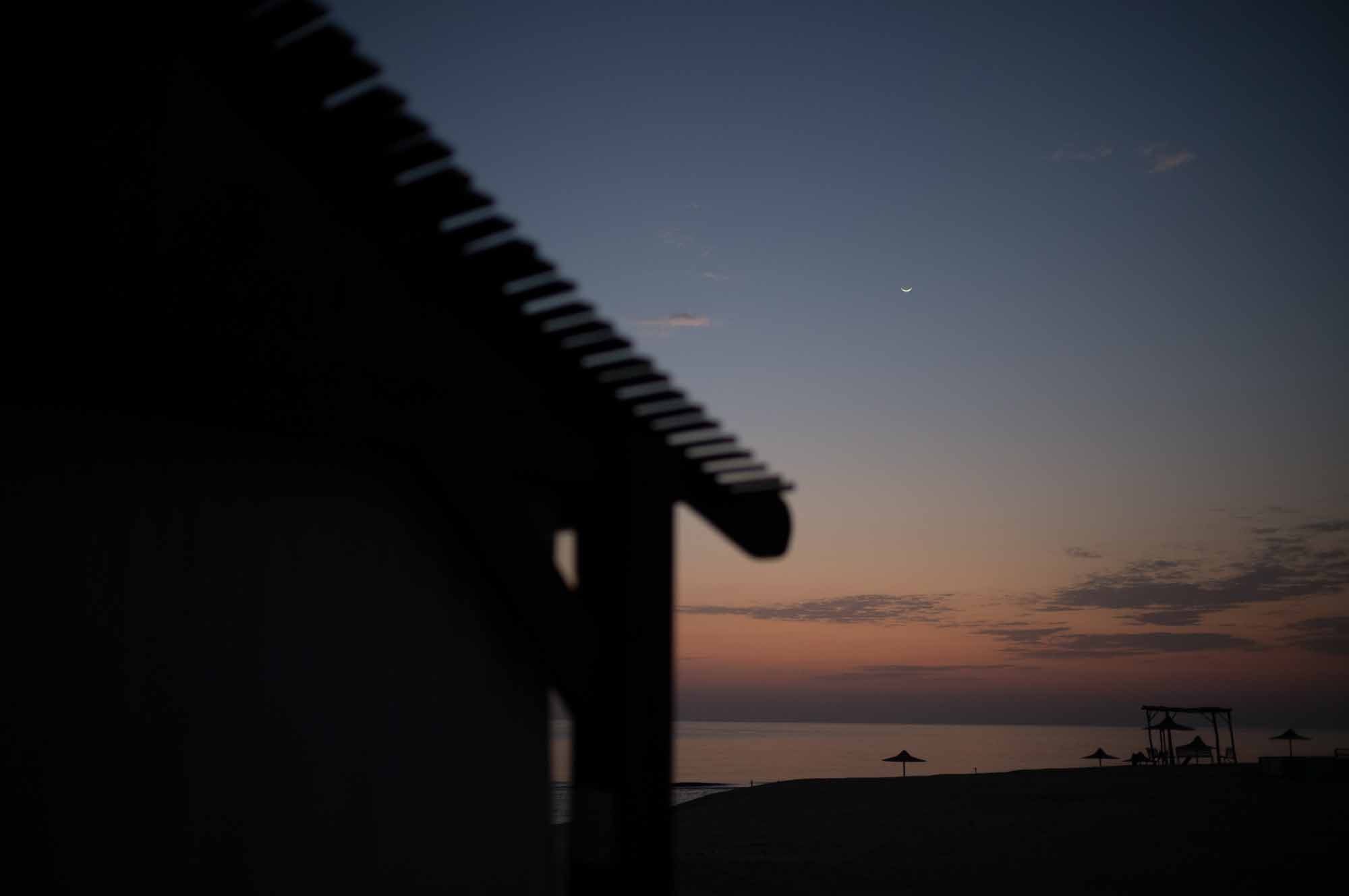 Sunset beach scene with silhouette of a hut, calm sea, crescent moon, and parasols on the horizon.