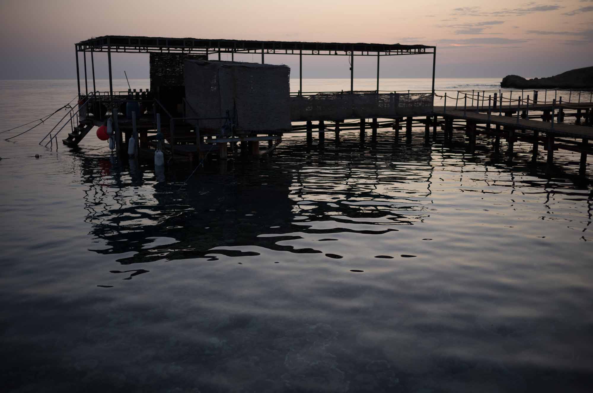 Serene ocean dock at sunset with calm water reflections and a silhouetted structure.