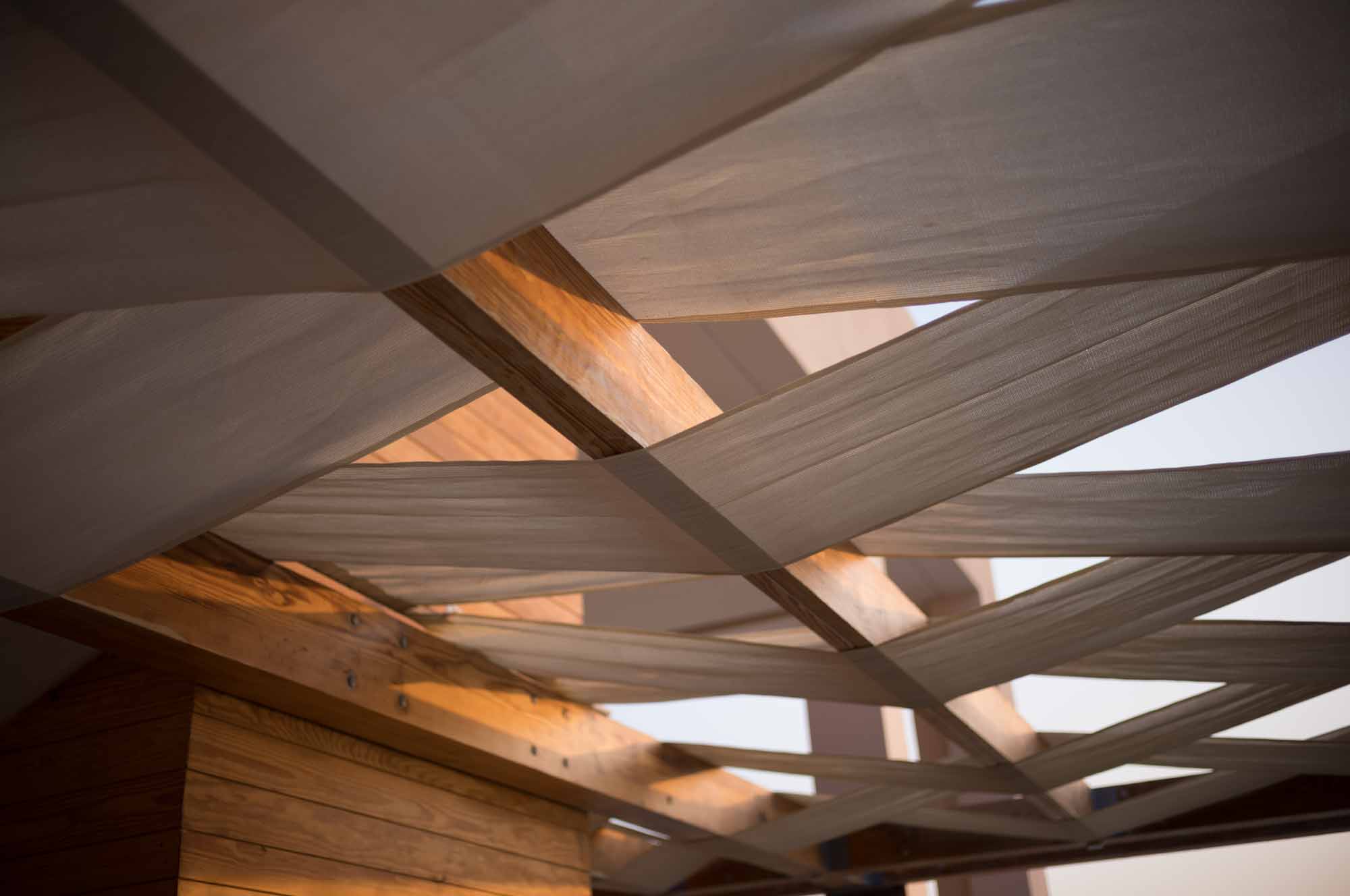 Modern wooden pergola with overlapping fabric panels creating shade and geometric design.