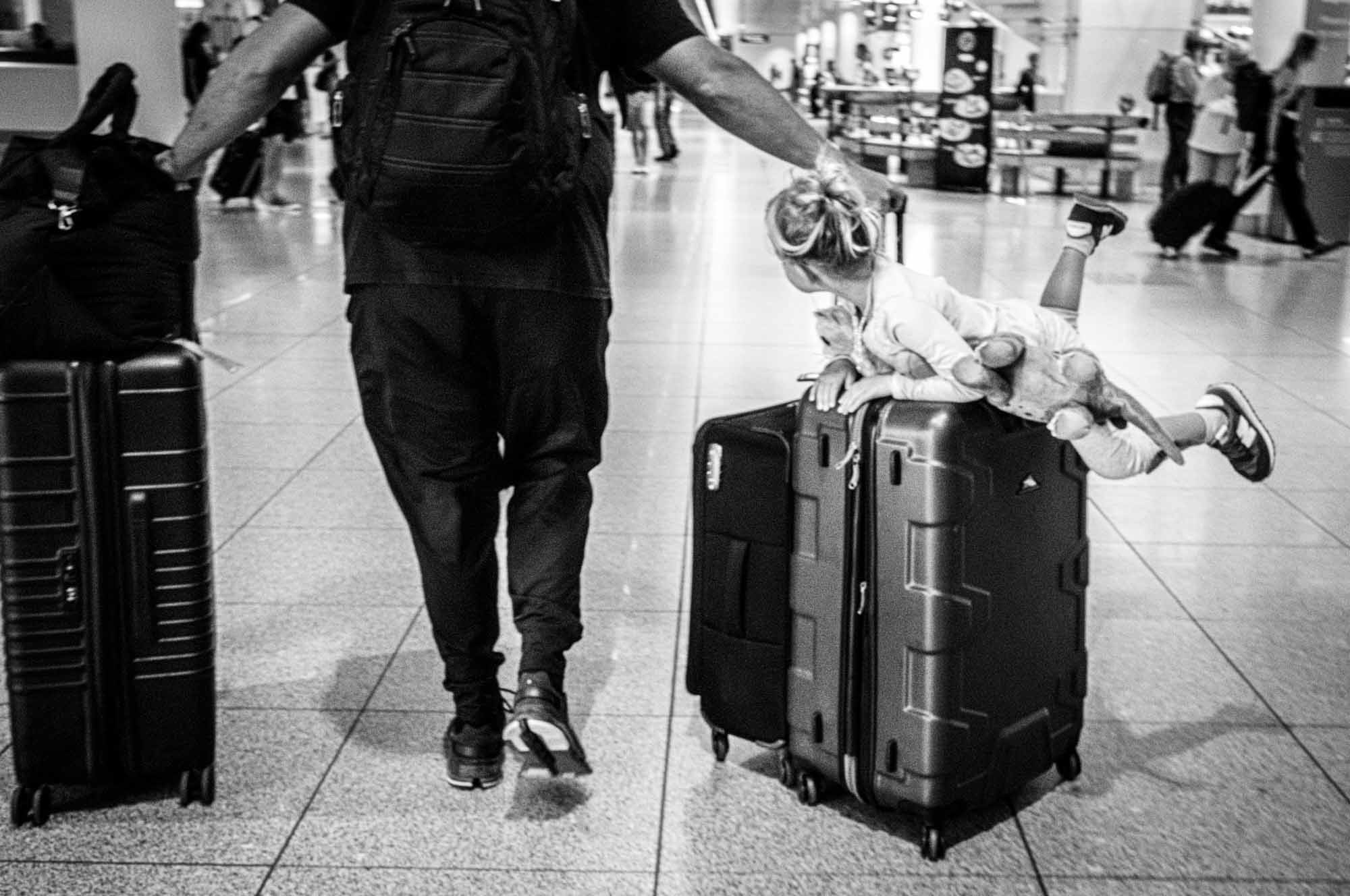 Man with luggage pulls child riding suitcase in busy airport terminal.