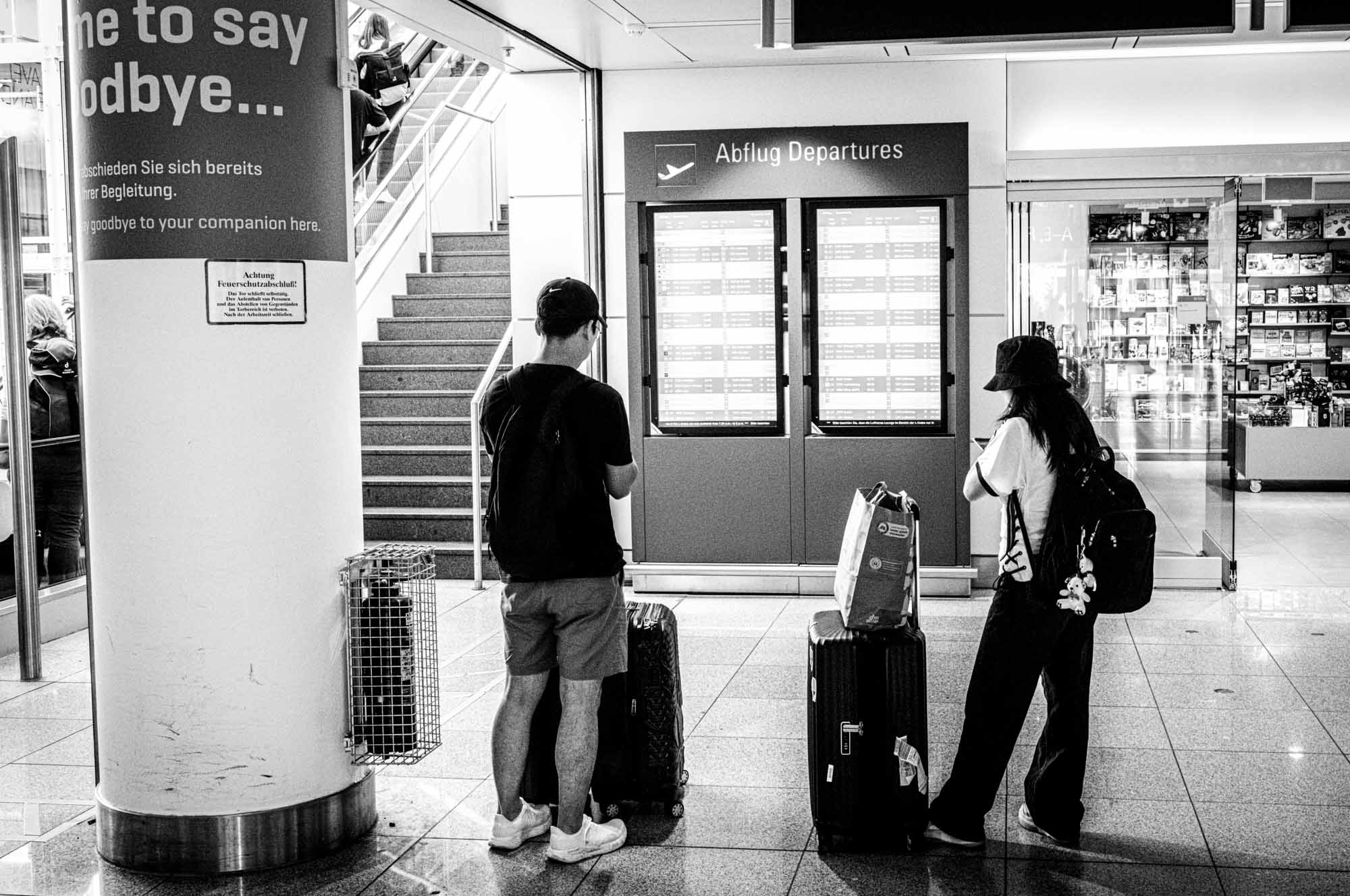 Travelers checking departure board at airport terminal, luggage beside, in black and white.