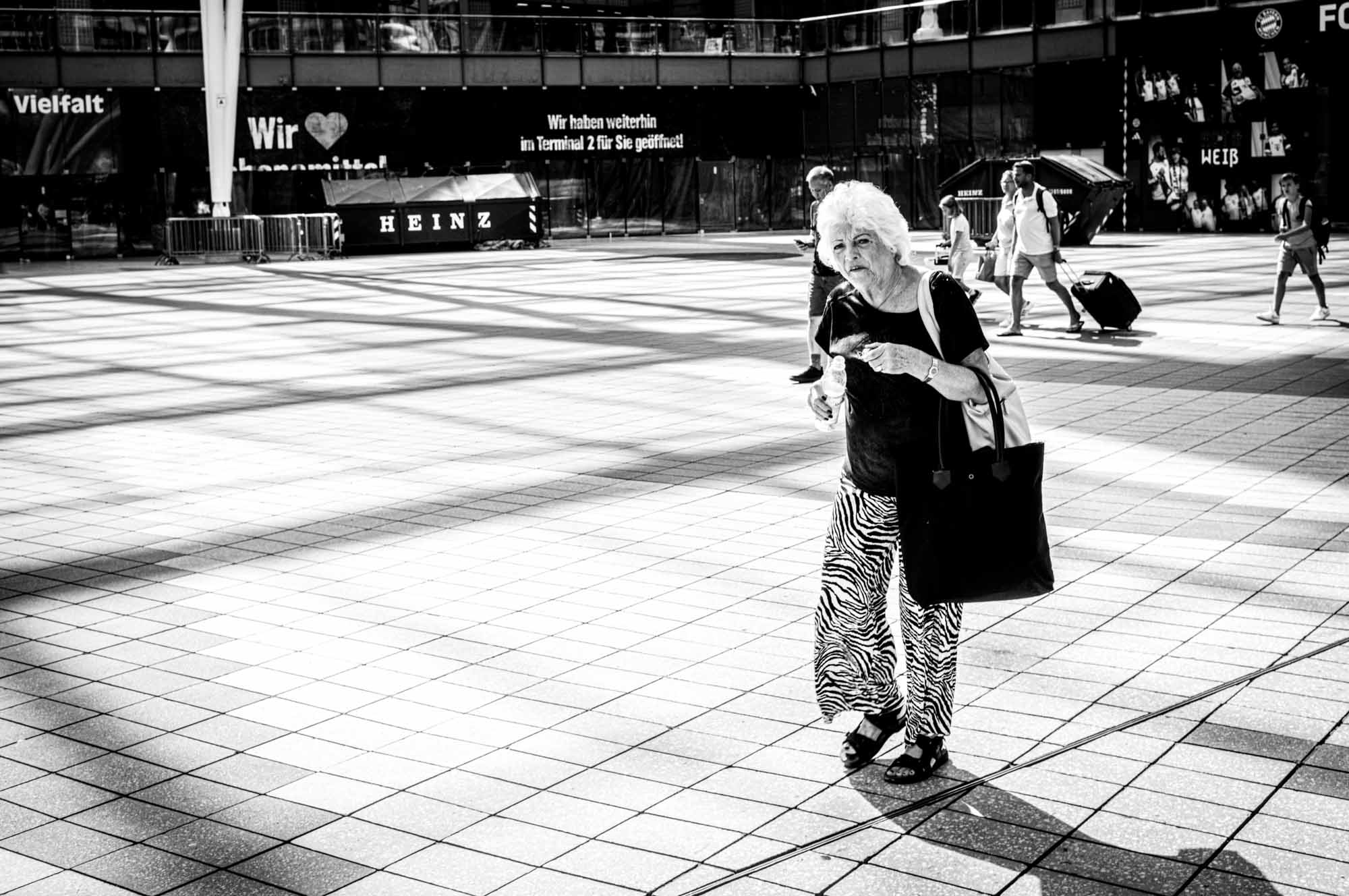 Elderly woman walking in a plaza with a tote bag, zebra-print pants, and shadows on the ground. Black and white.