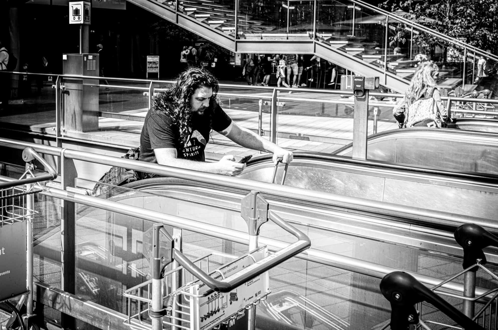 Man with long hair using phone on escalator at shopping center in black and white.