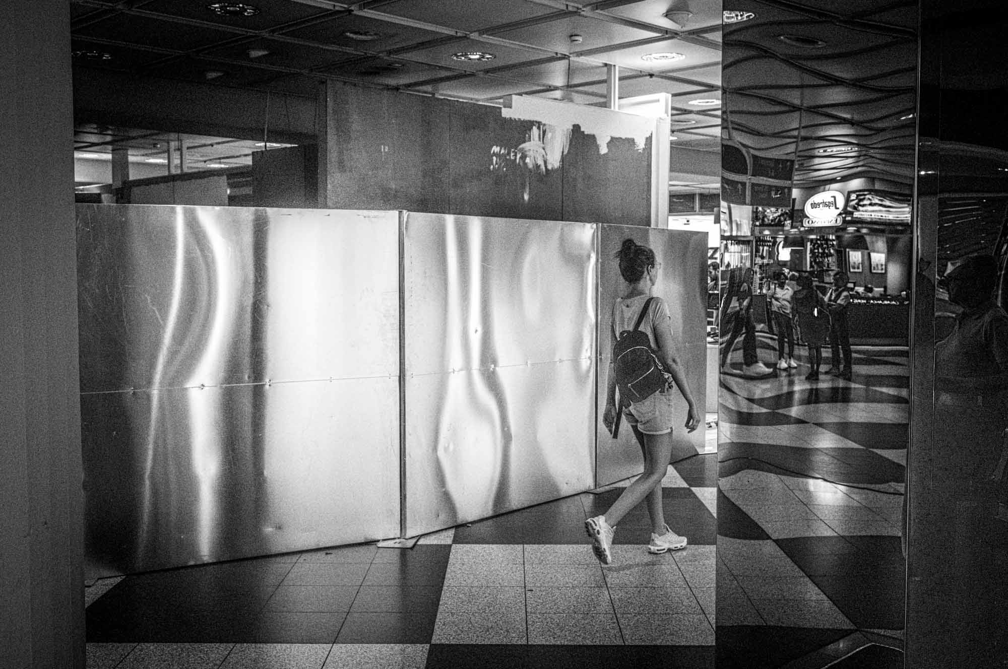 Woman with backpack walks past metal barrier inside a dimly lit shopping mall with checkered floors.