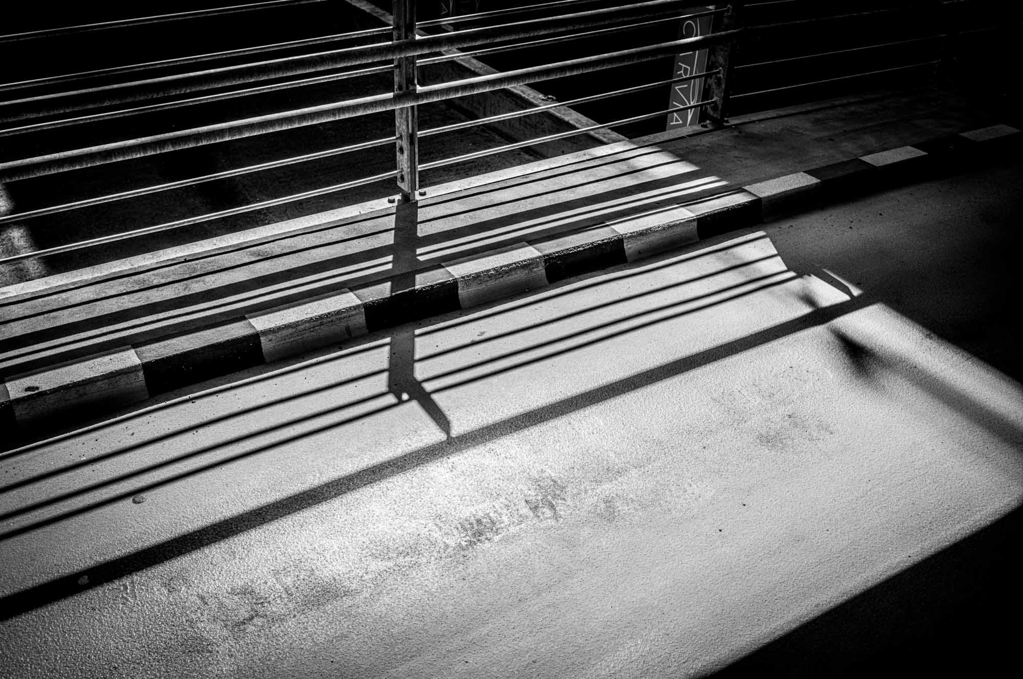 Black and white shadows cast by railings on pavement, creating geometric patterns in sunlight.