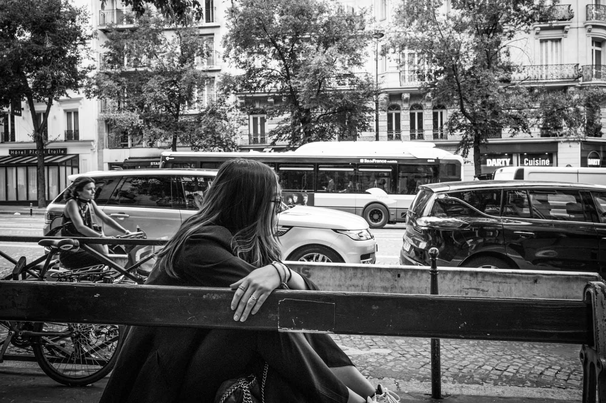 Woman sitting on a bench in the city, watching traffic, with a cyclist passing by. Black and white urban scene.