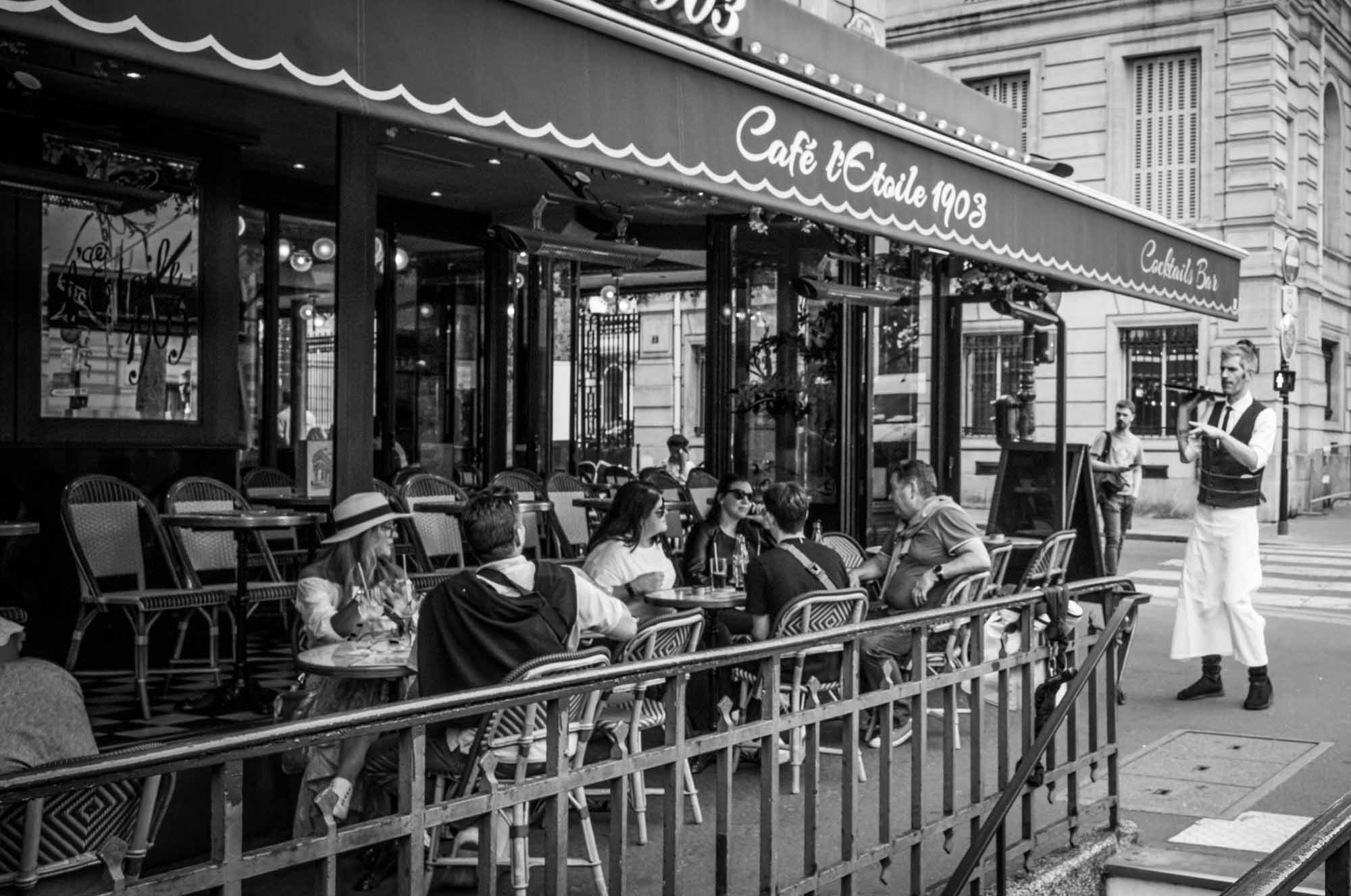 Outdoor seating at a bustling Parisian cafe with people dining and a waiter serving in black and white.