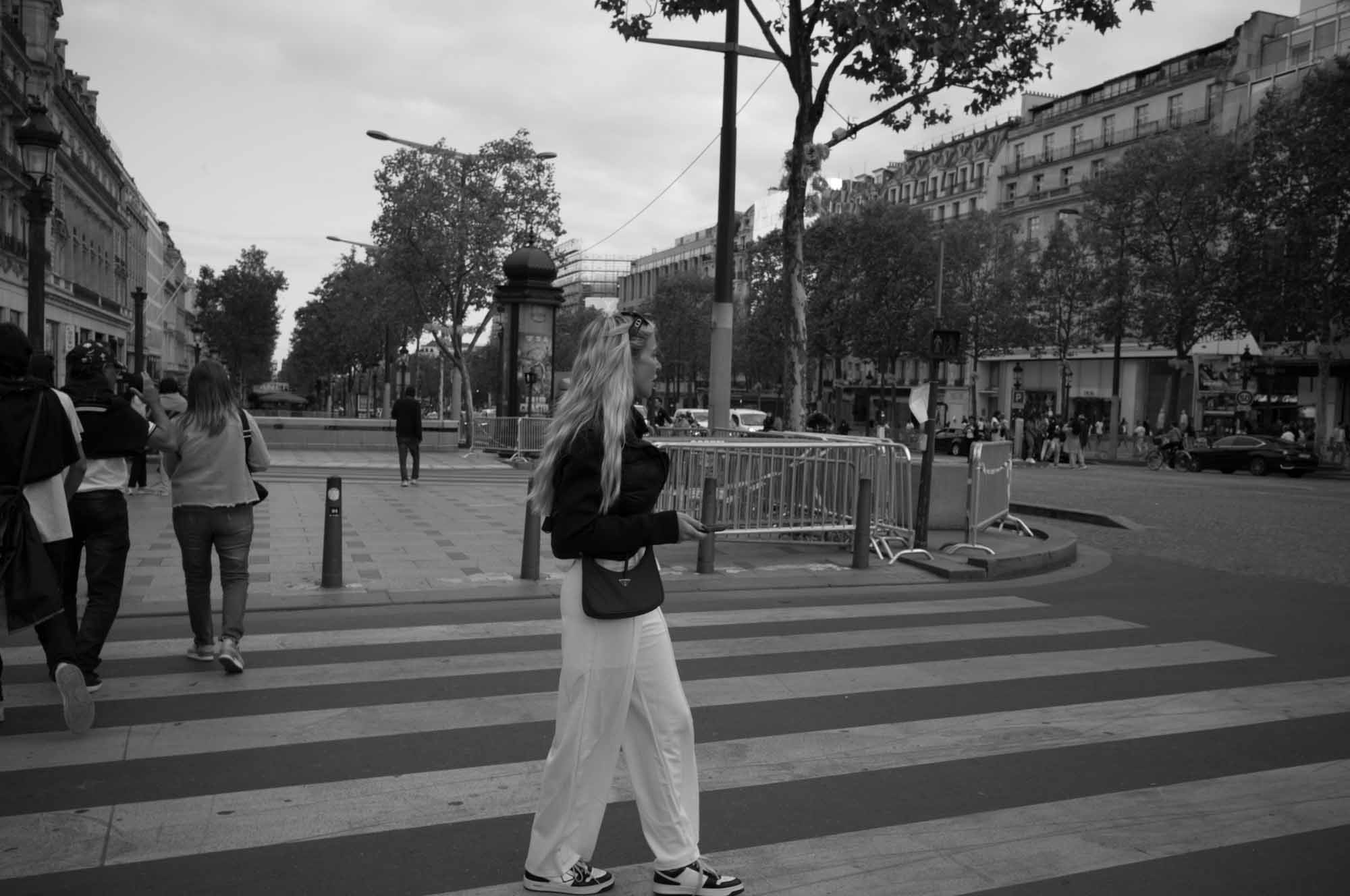 Woman crossing a city street with striped pedestrian crosswalk and trees lining the road.