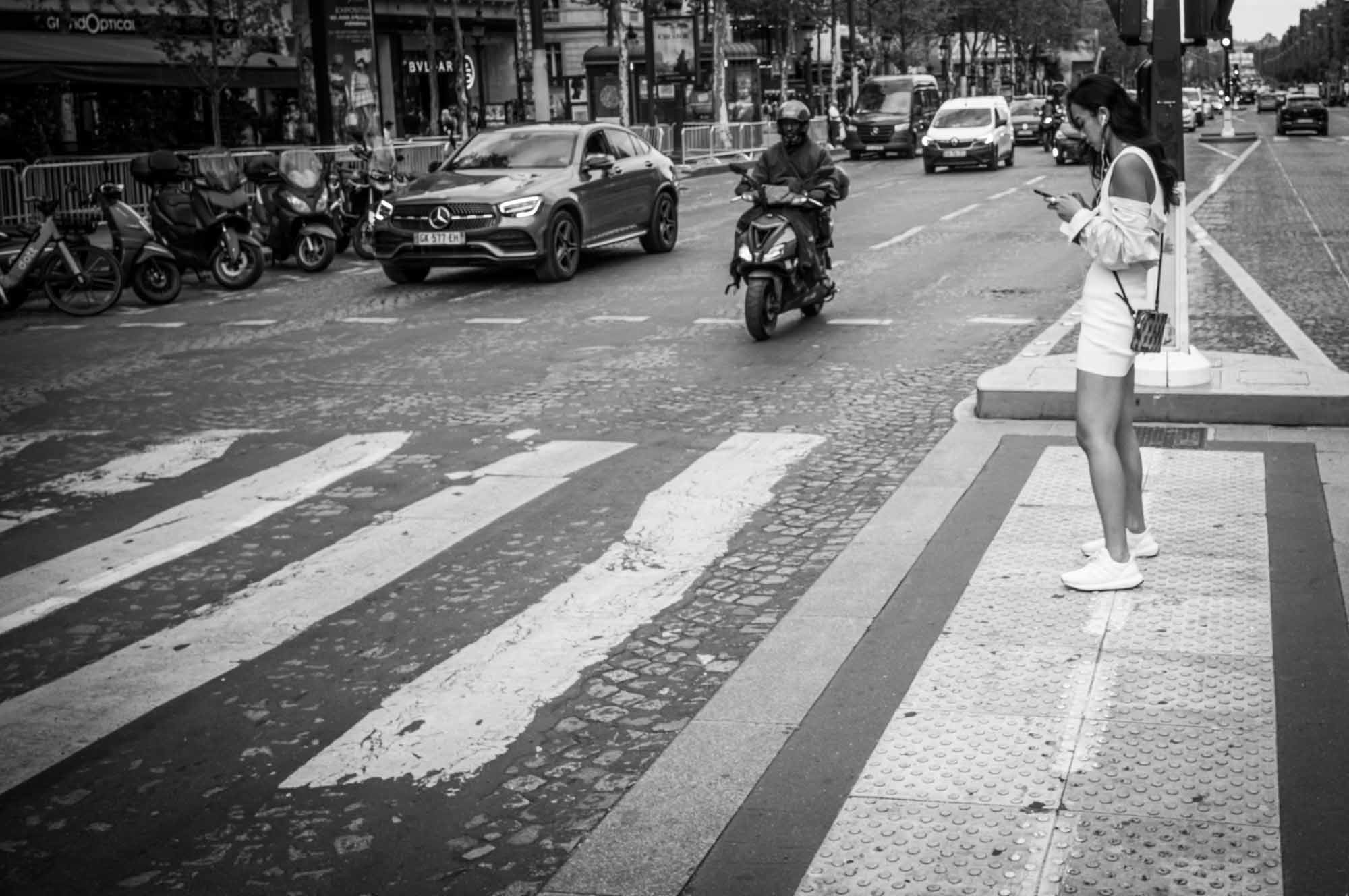 Woman stands at pedestrian crossing on busy city road, checking phone. Black and white street scene.