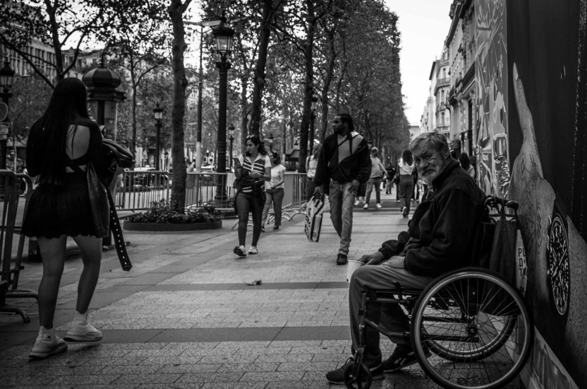 Black and white street scene with people walking and a man in a wheelchair on a city sidewalk.