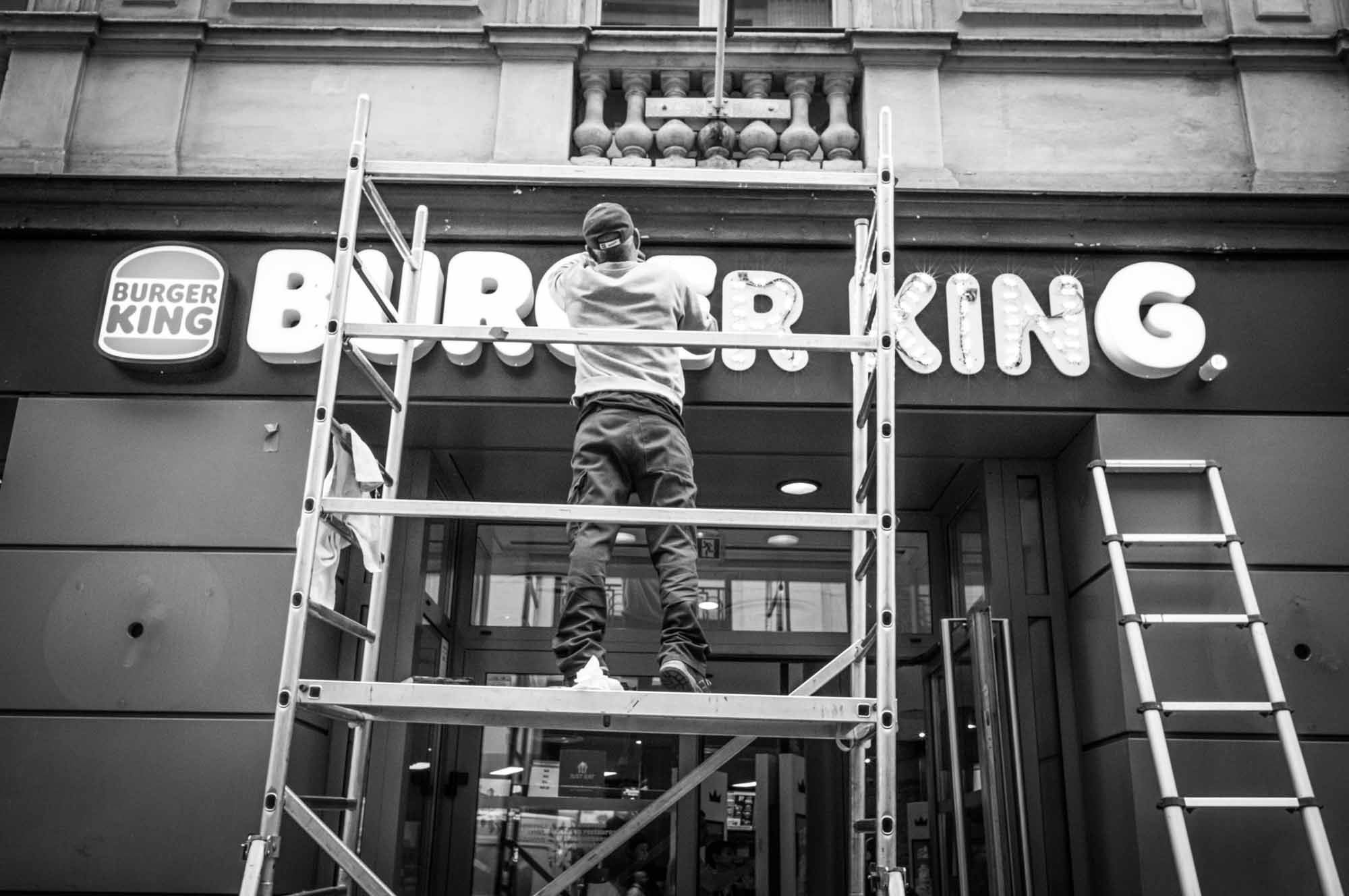 Worker on scaffolding repairing Burger King sign in black and white street scene.