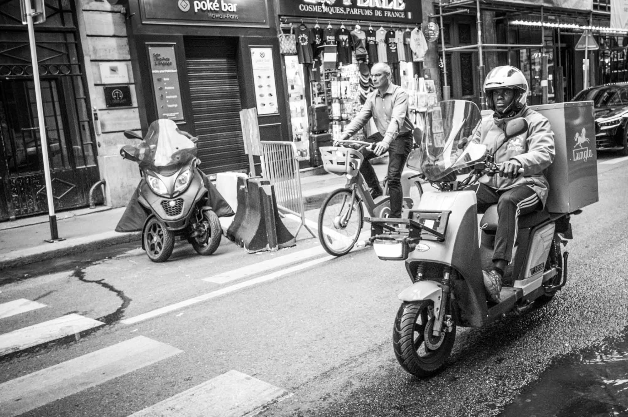 Delivery scooter and cyclist on a bustling city street near a poke bar. Black and white urban scene.