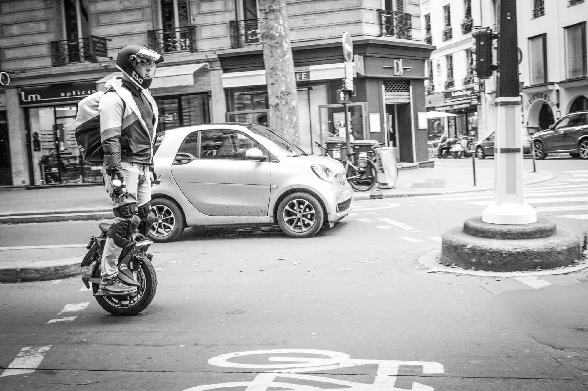 Person riding an electric unicycle on a city street, wearing helmet and gear, next to parked cars and bicycles. Black and white.