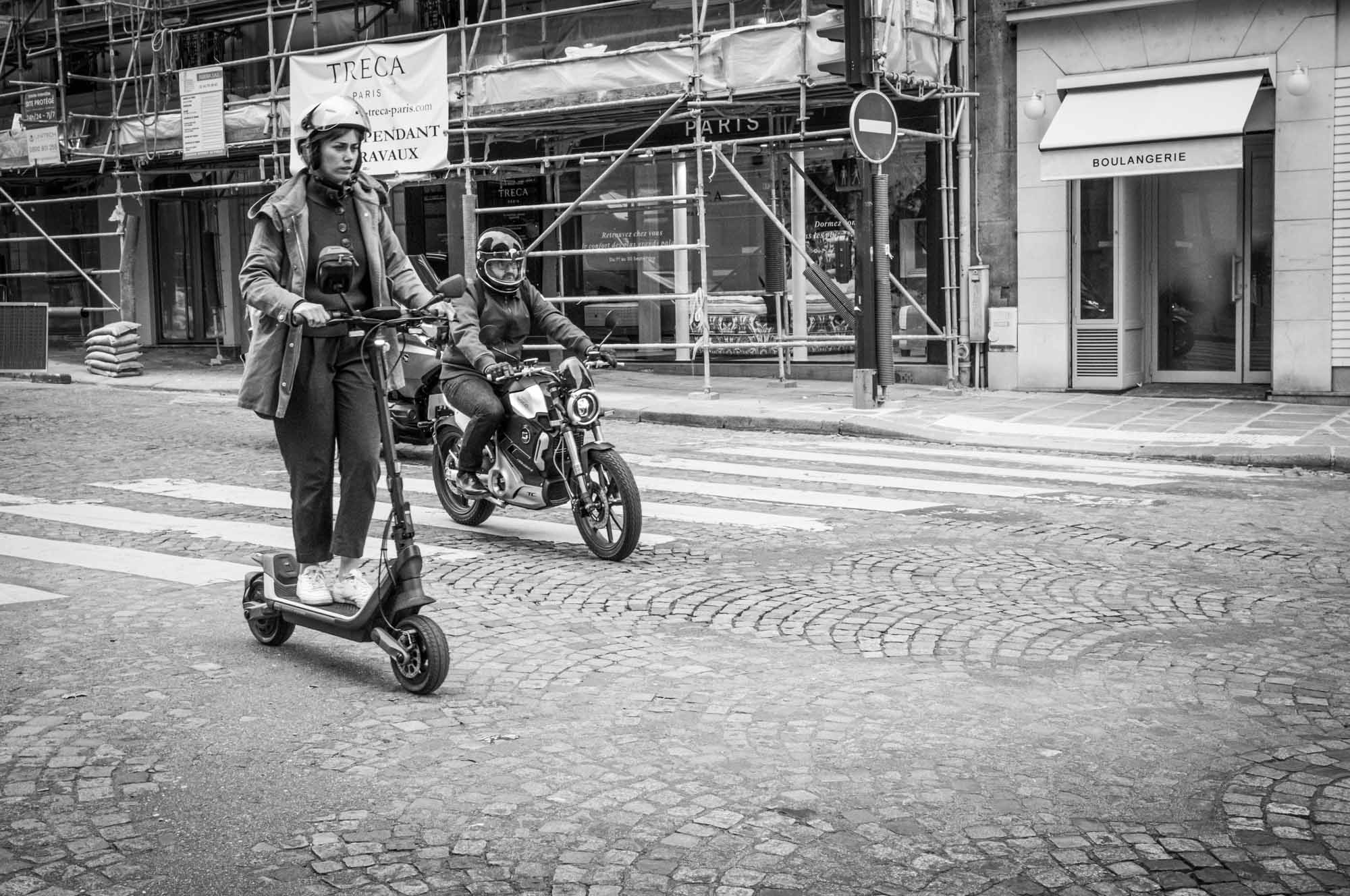 Person rides an electric scooter beside a motorbike on a cobblestone street, city backdrop.