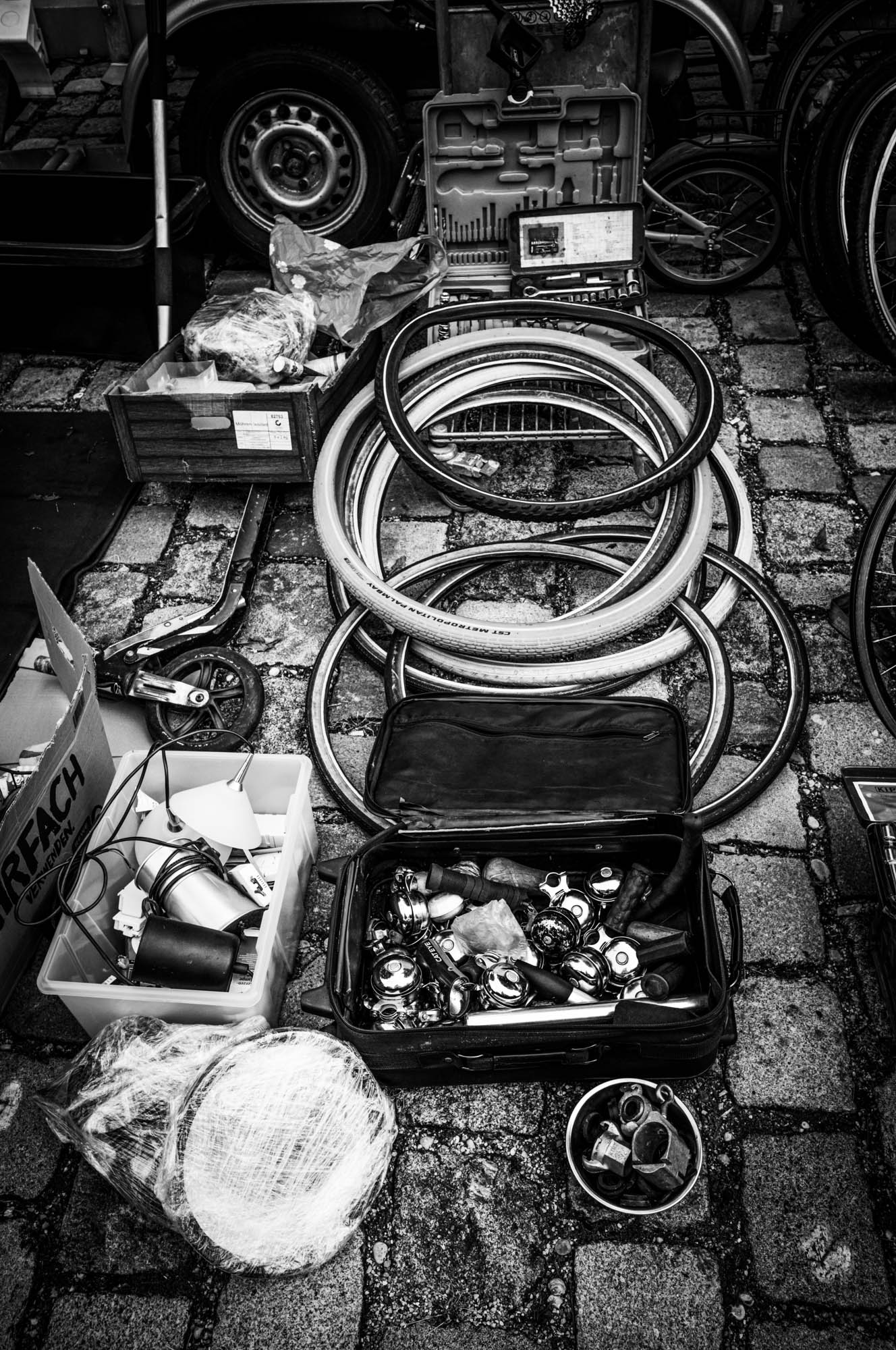 Flea market bicycle parts: tires, bells, and tools on cobblestone street, black and white photography.