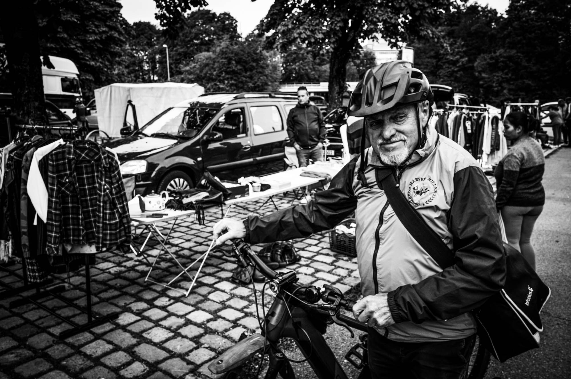 Elderly man wearing a helmet and jacket stands with a bike at an outdoor market, surrounded by clothes and people.