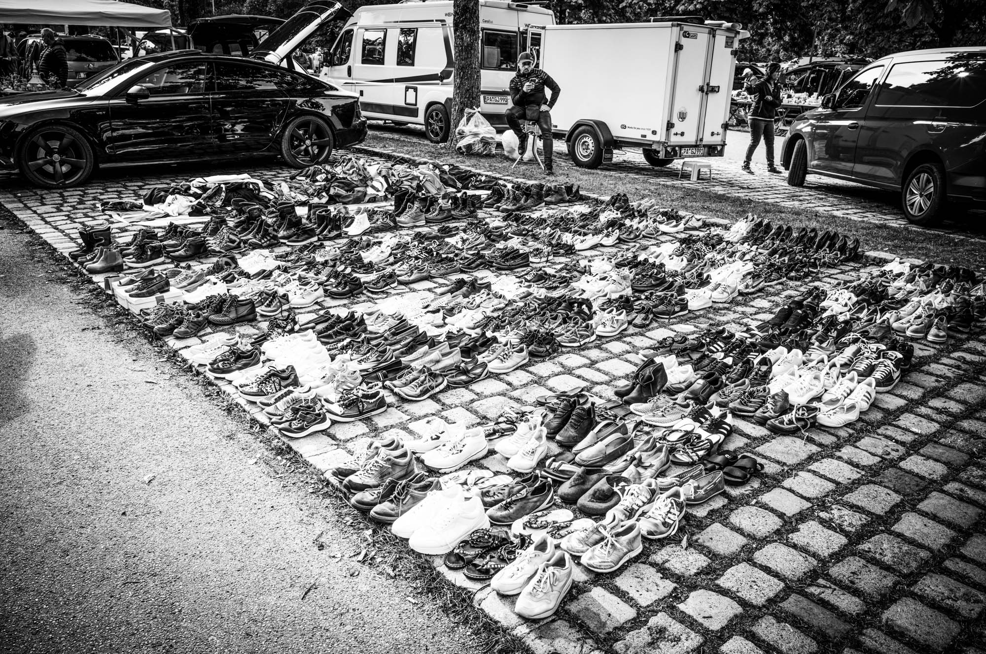 Outdoor market with rows of shoes on pavement near cars; monochrome photo of a street sale.