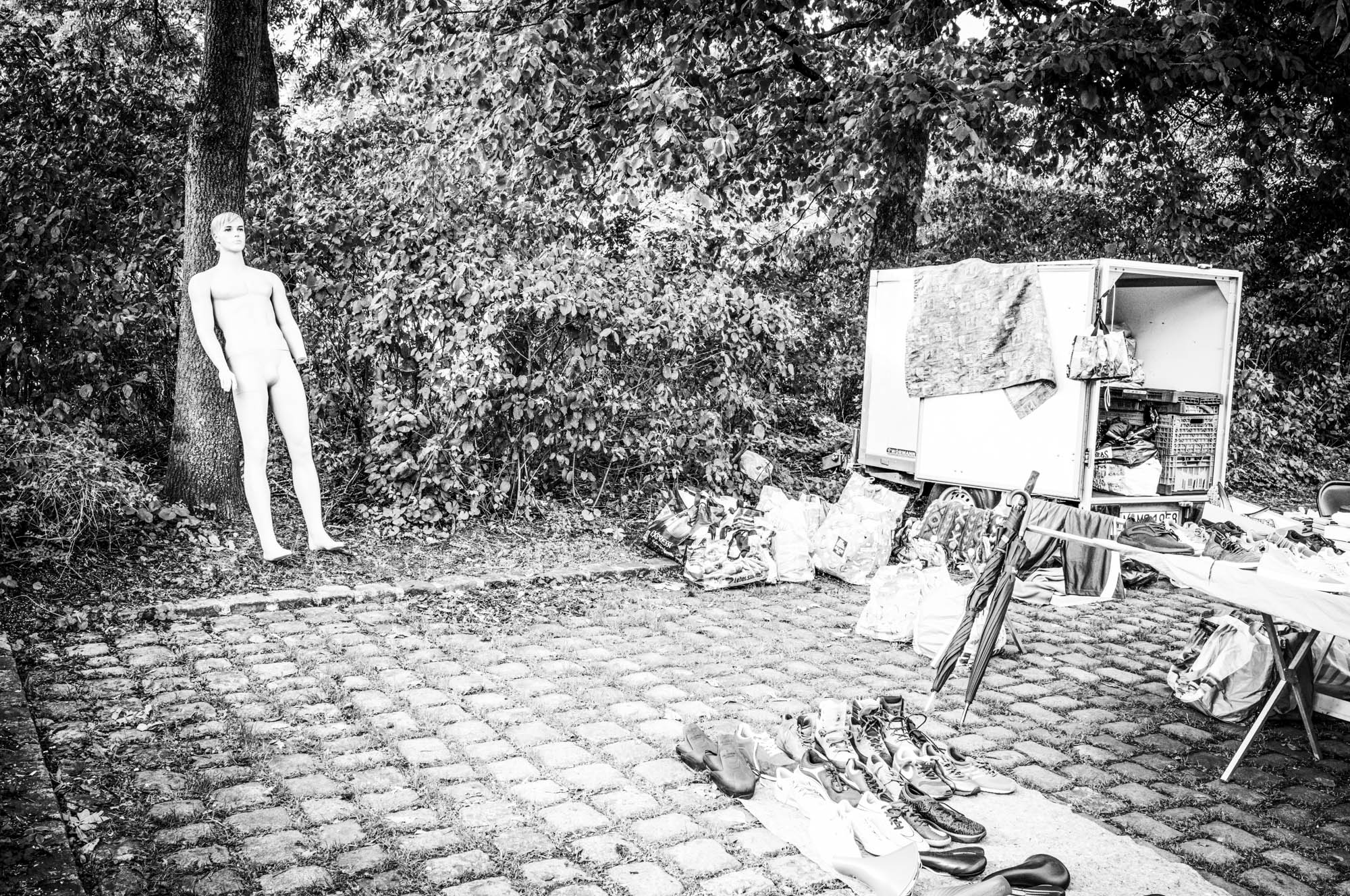 Mannequin near a tree in an outdoor flea market setting, surrounded by assorted goods and cobblestone pavement.