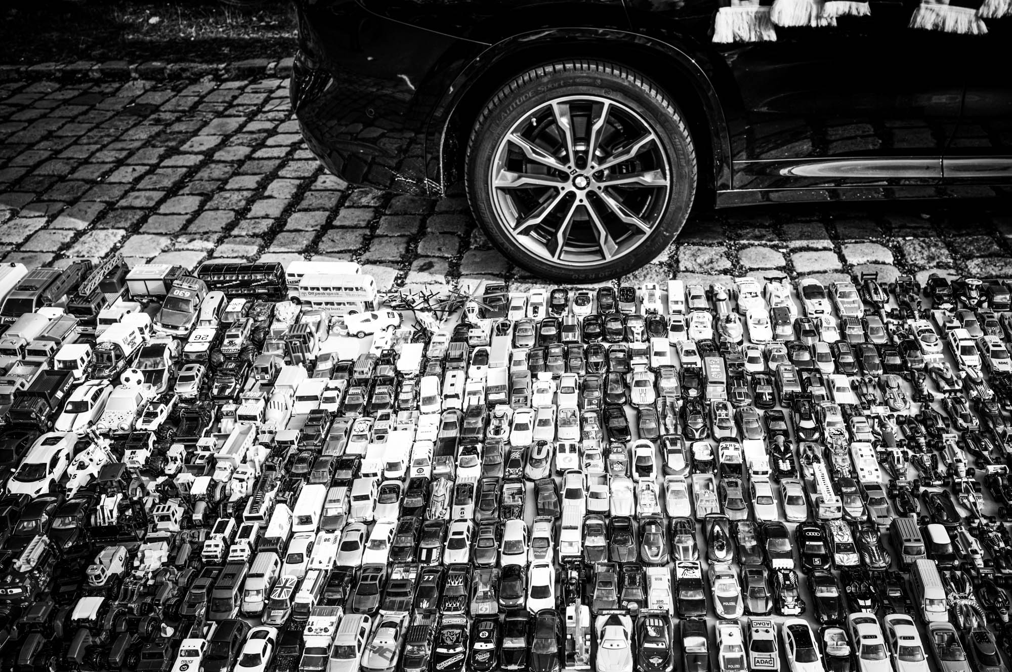 Black and white photo of a car wheel next to rows of toy cars on cobblestones.