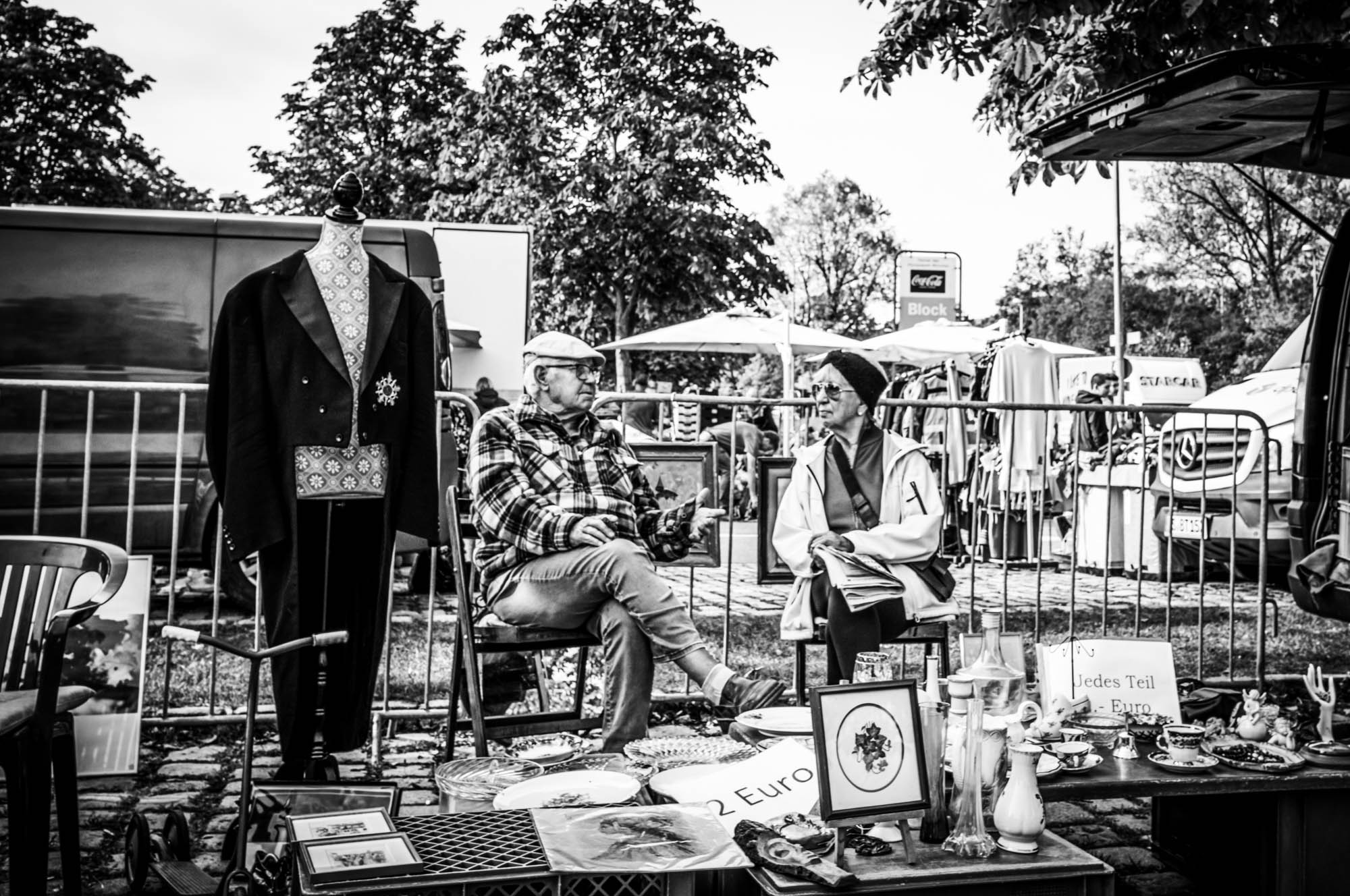 Vintage market scene with two people chatting, surrounded by antiques and clothes in black and white.