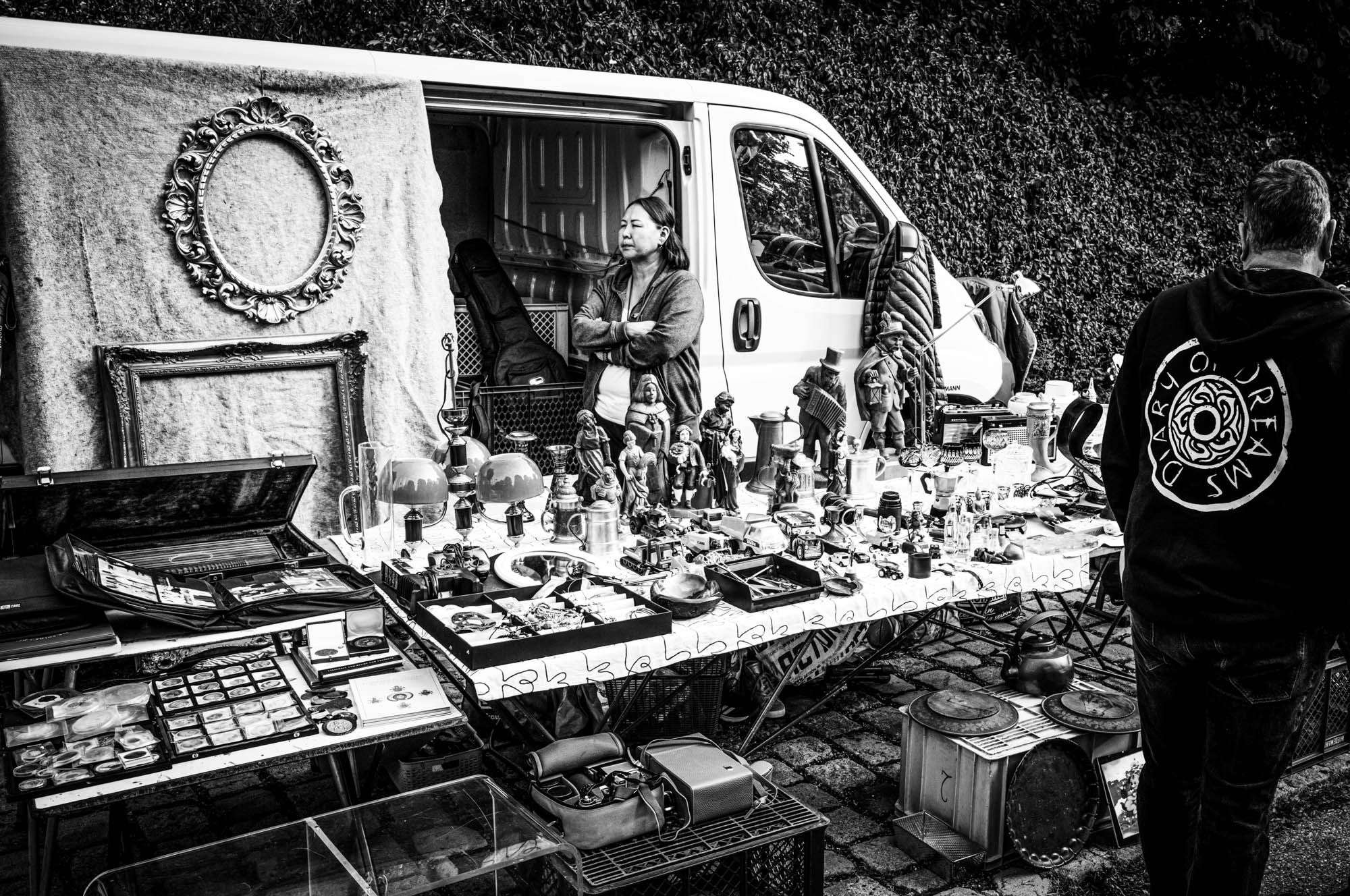 Black and white photo of a flea market stall with various vintage items and a vendor beside a white van.