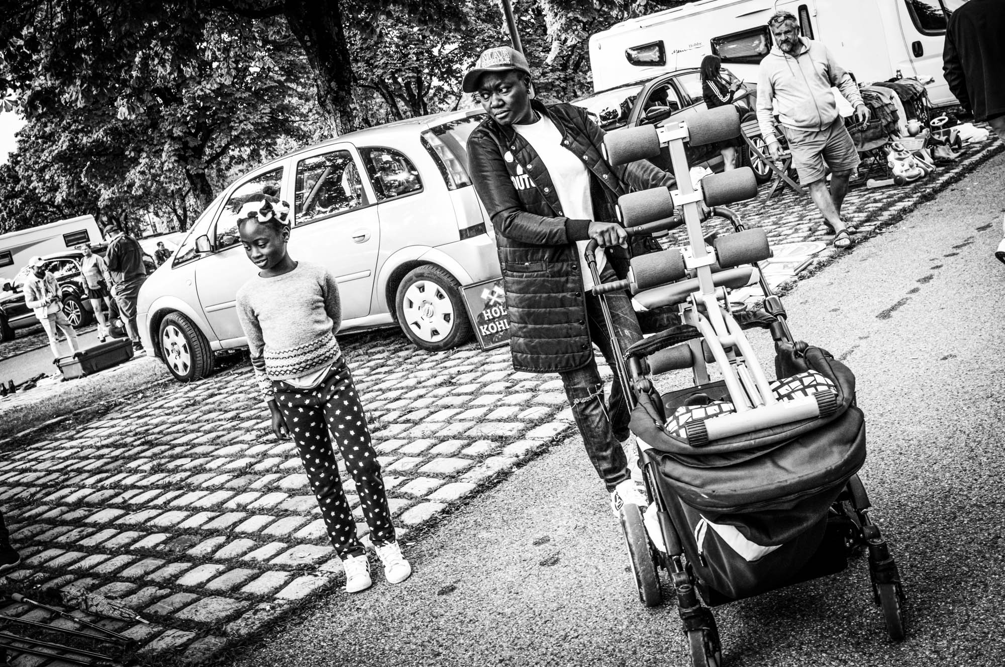 Black and white street scene: person with stroller, parked cars, and people in background on a cobblestone path.