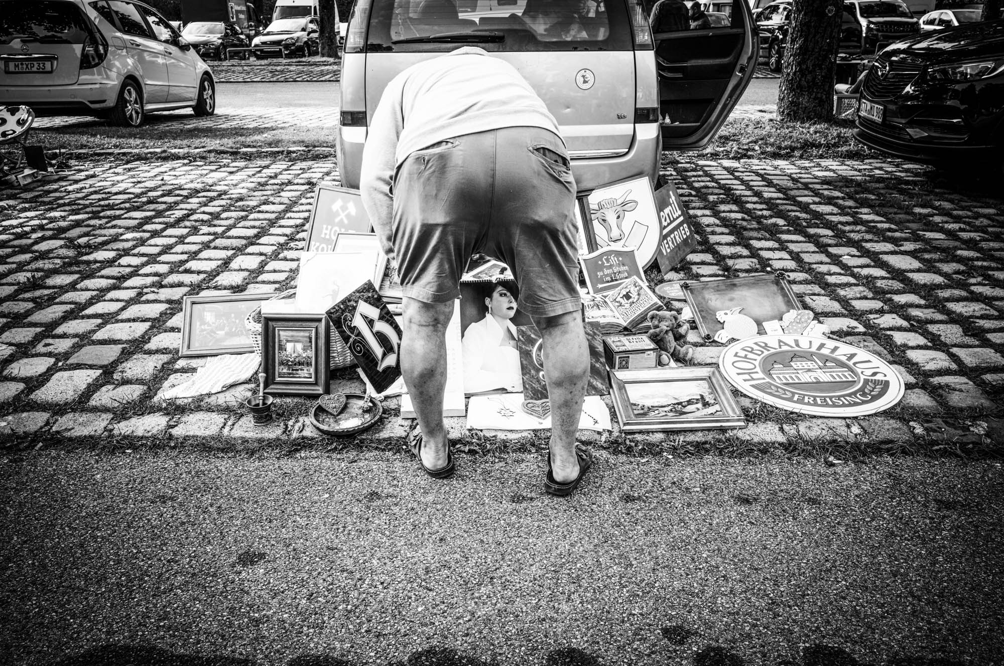 Man browsing vintage items for sale at a flea market beside a parked car. Black and white.