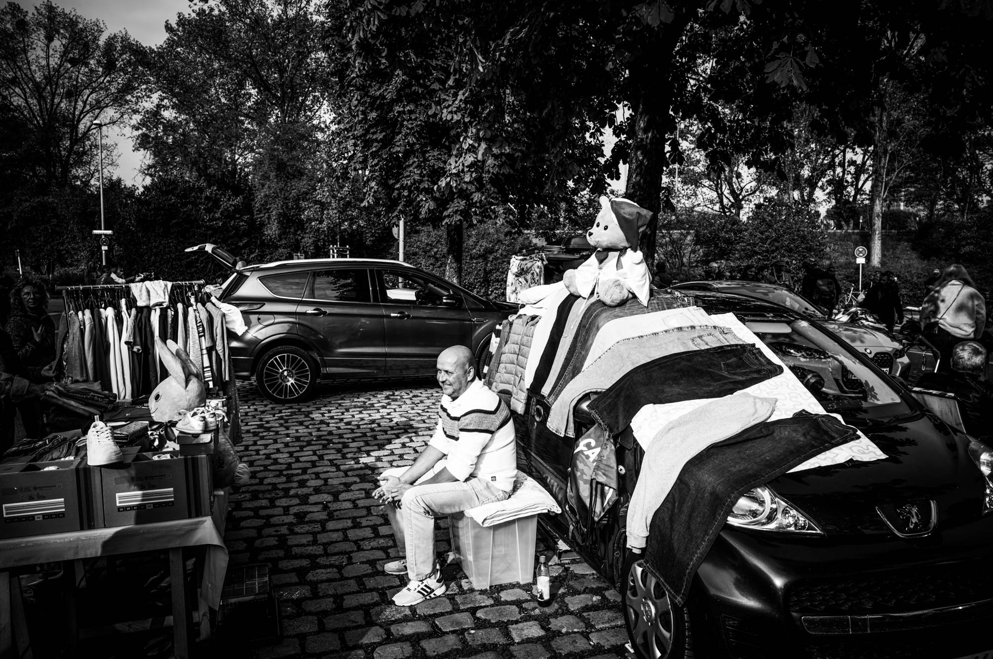 Man sitting at outdoor market stall with clothes and stuffed toy on cars, black and white photo.