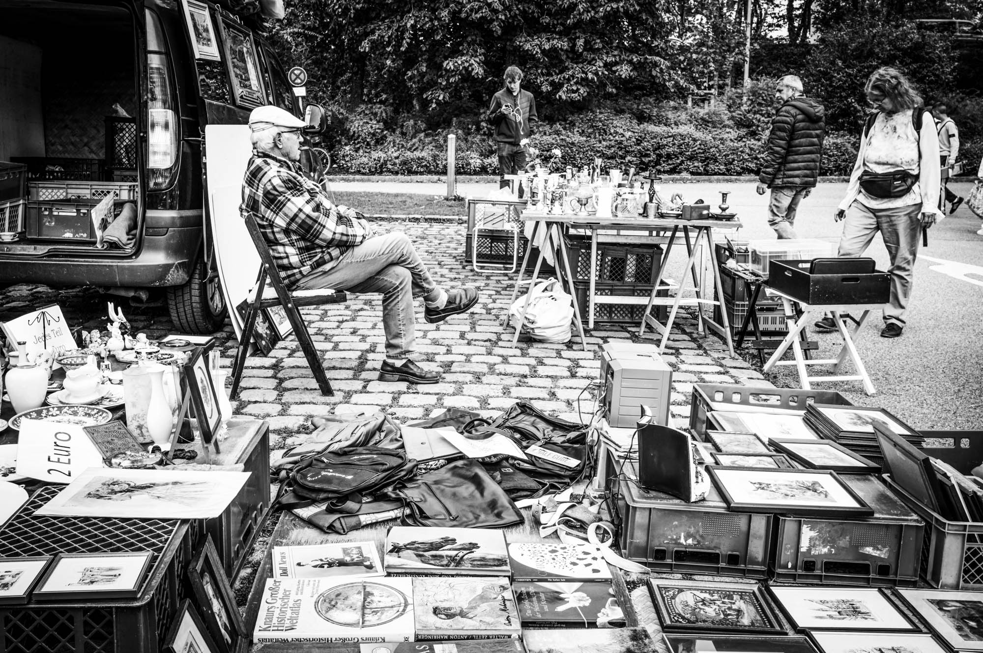 Black and white photo of a man sitting by his flea market stall with various items on display.