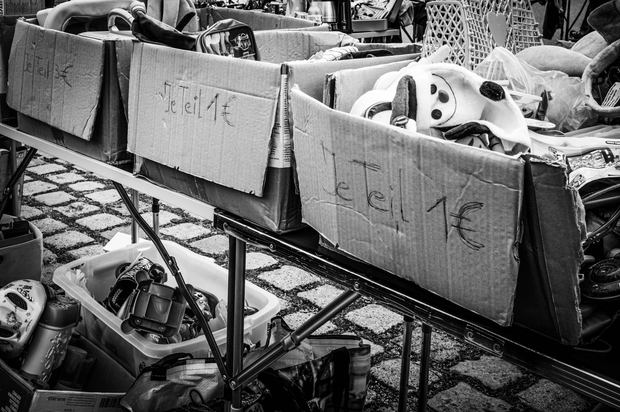 Cardboard boxes at a flea market labeled Je Teil 1β¬, filled with assorted items for sale.