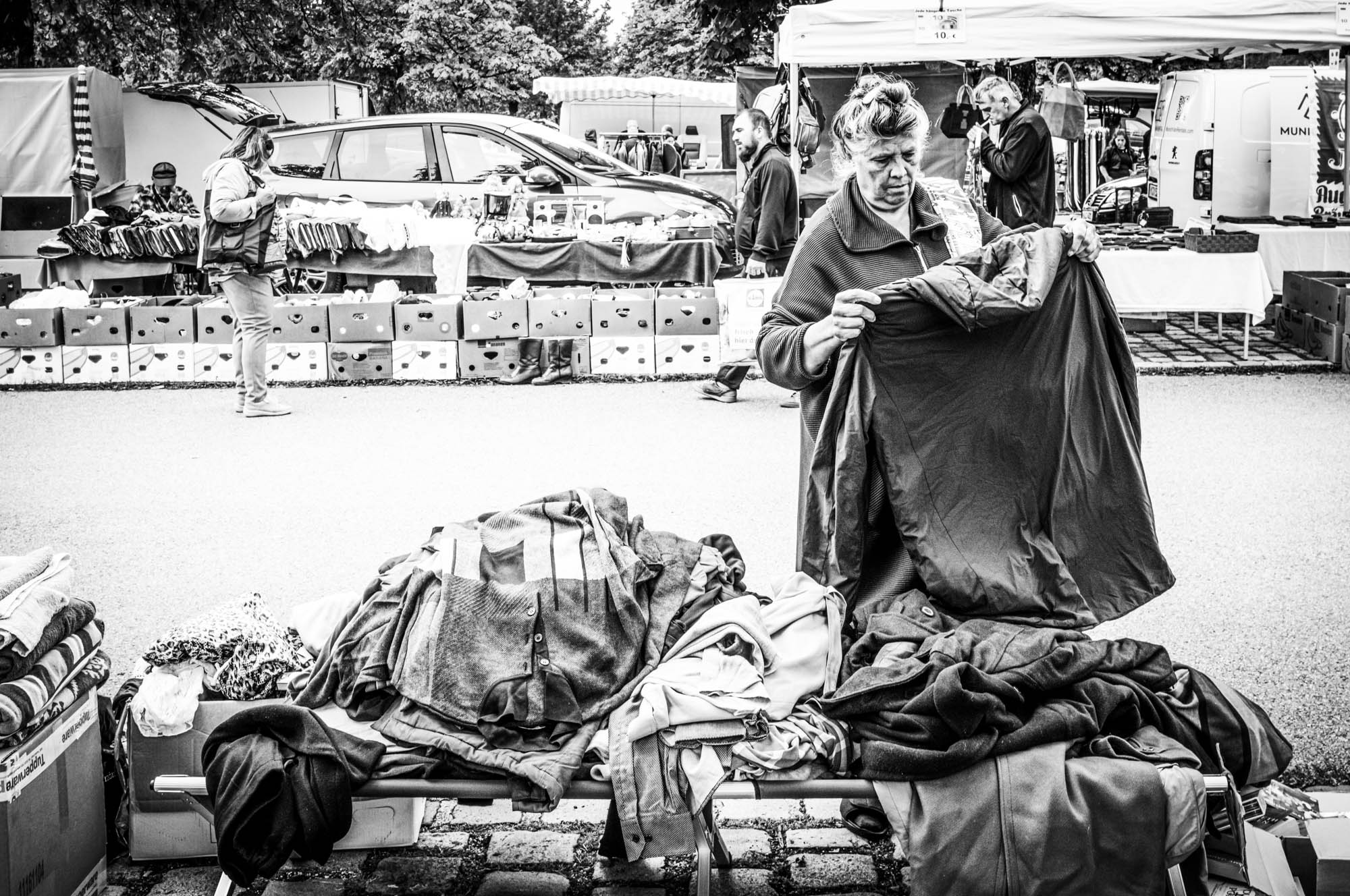 Market scene with woman examining clothes; stalls and shoppers in the background. Black and white photograph.