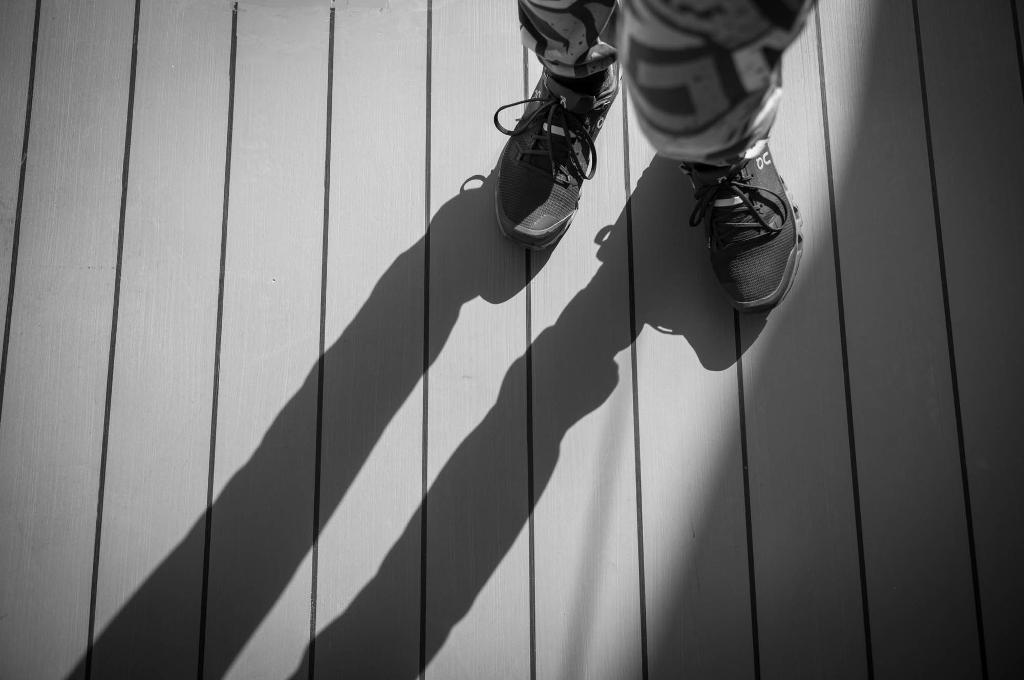 Black and white photo of person in sneakers casting long shadows on wooden deck.