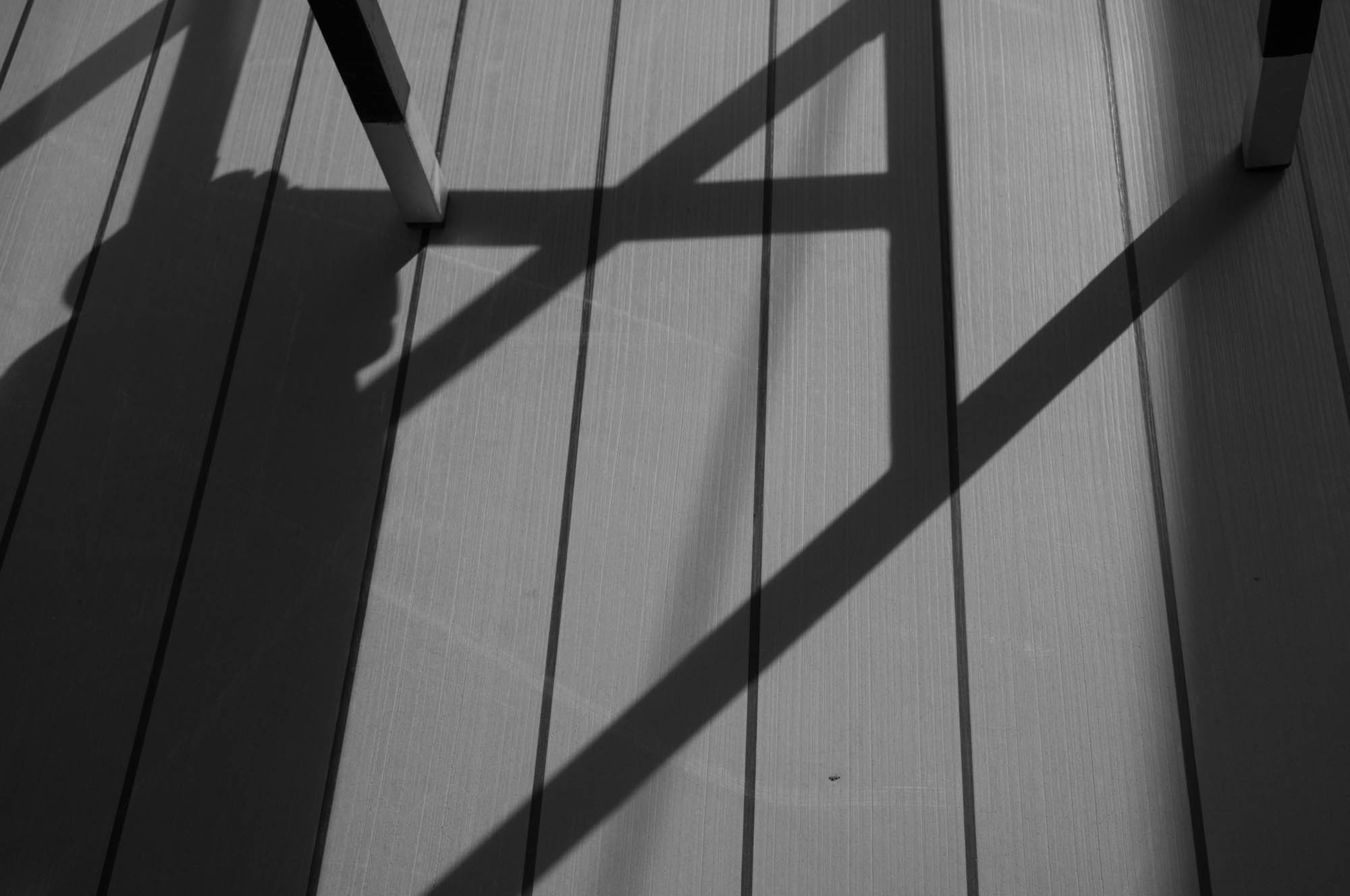 Shadow of a chair on wooden deck boards, creating geometric pattern in sunlight, black and white image.