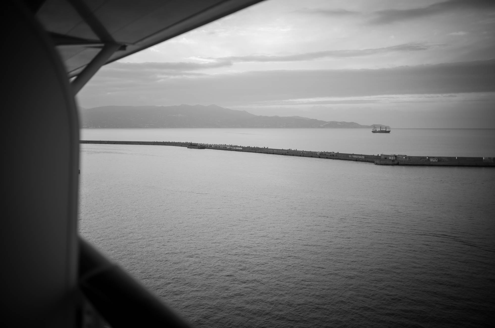 Black and white view of a serene ocean, distant mountains, and a ship near a pier under a cloudy sky.
