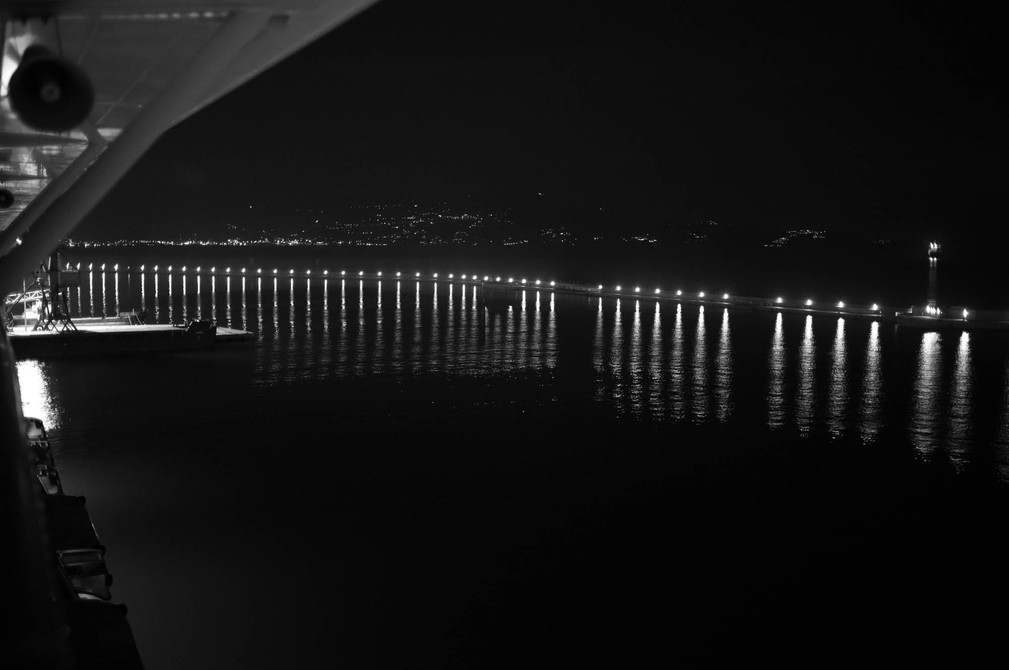 Nighttime harbor with lit pier and reflections on water. City lights in background create a serene waterfront view.