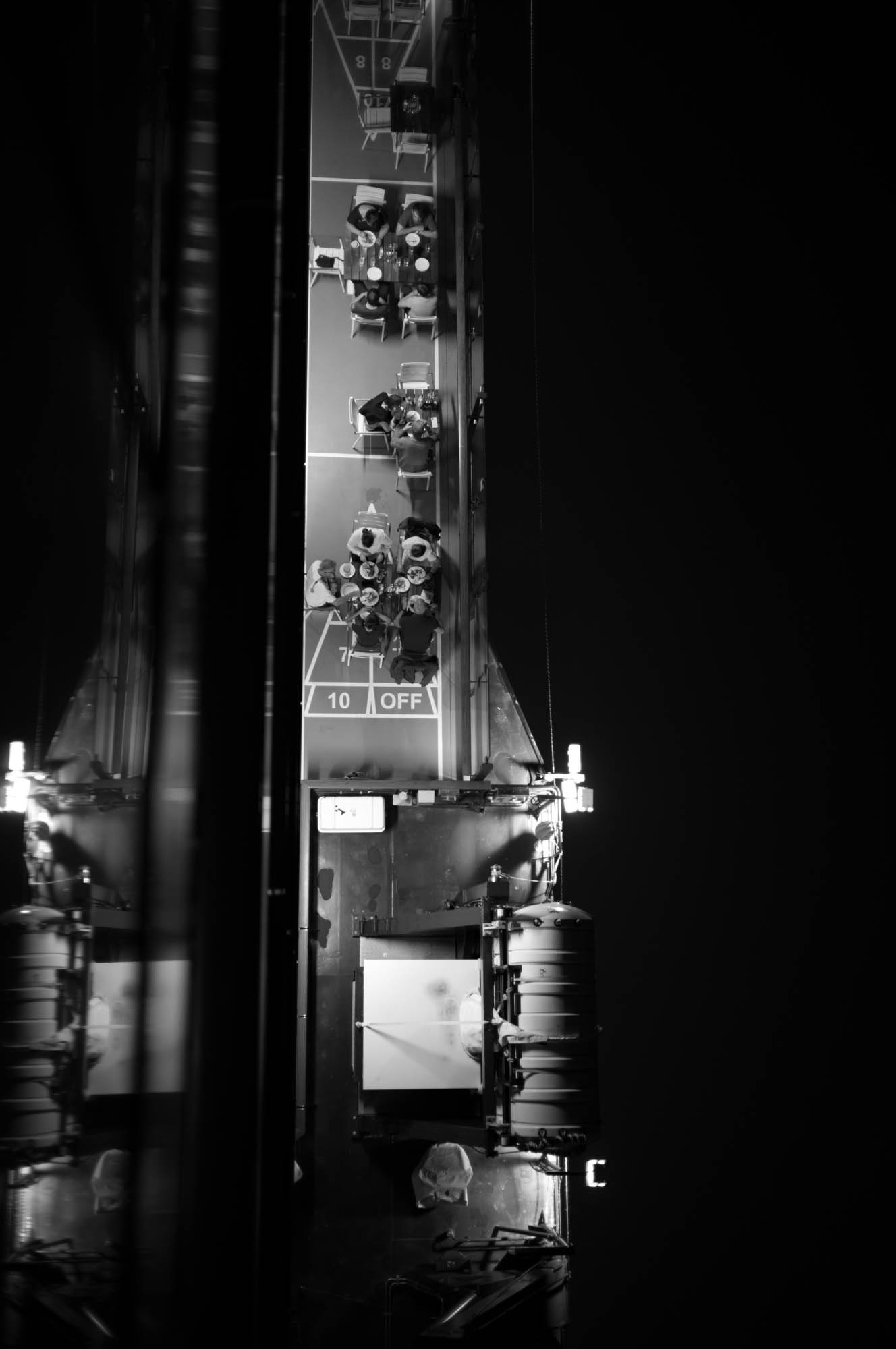 Aerial view of a dark ship deck with people seated at tables under bright lighting, captured at night.