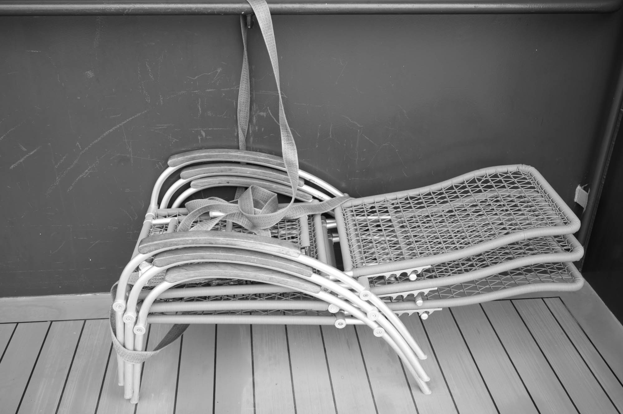 Stacked woven chairs secured with a strap, against a scratched wall, on a wooden deck. Black and white photo.