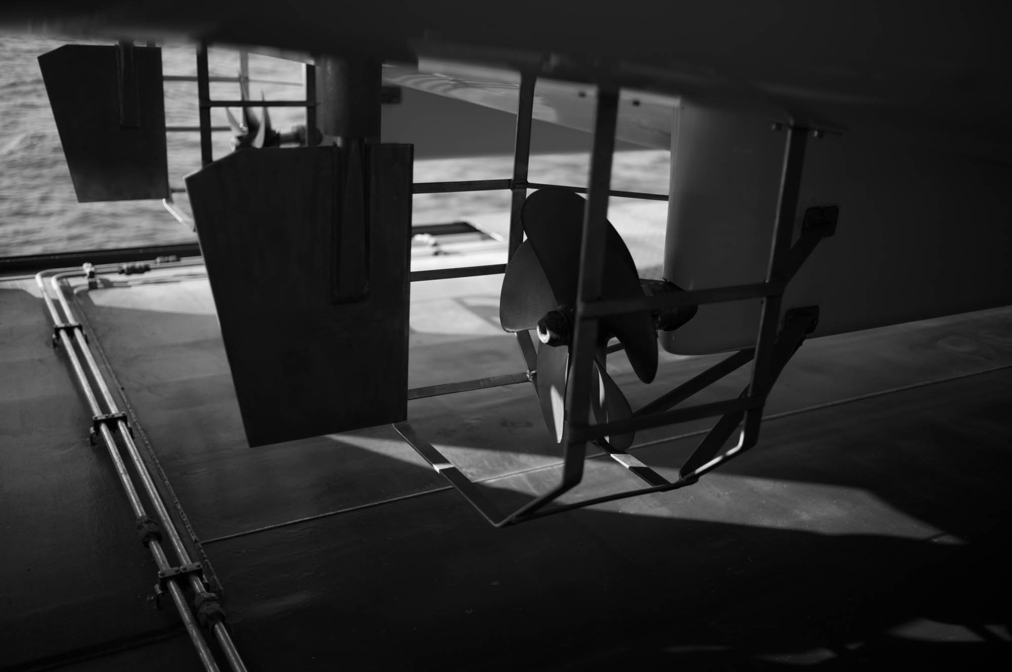 Black and white image of a boat's propeller and rudders under the deck, highlighting marine engineering components.