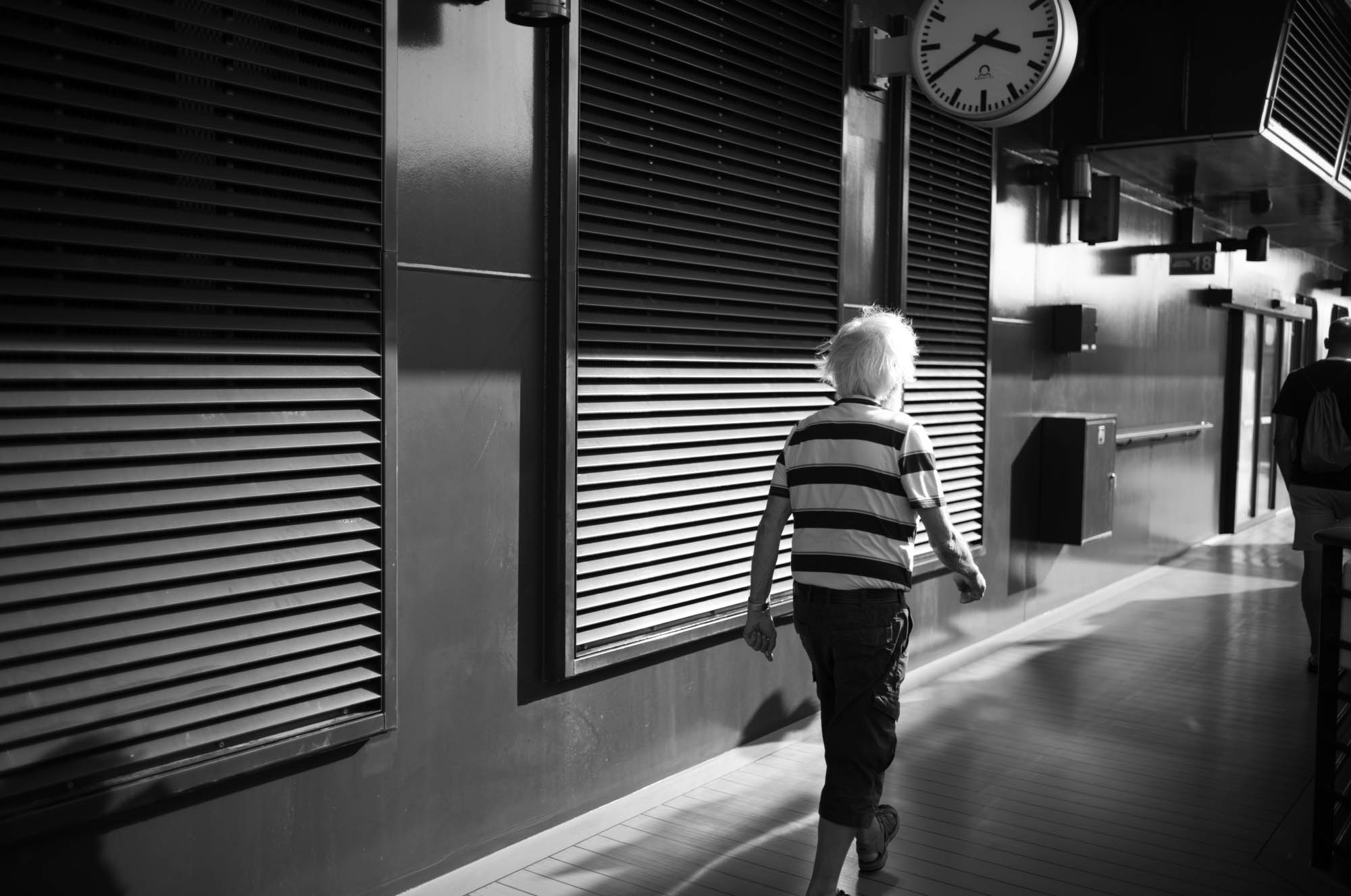 Elderly man in striped shirt walking through an empty urban corridor under a large clock, in black and white.