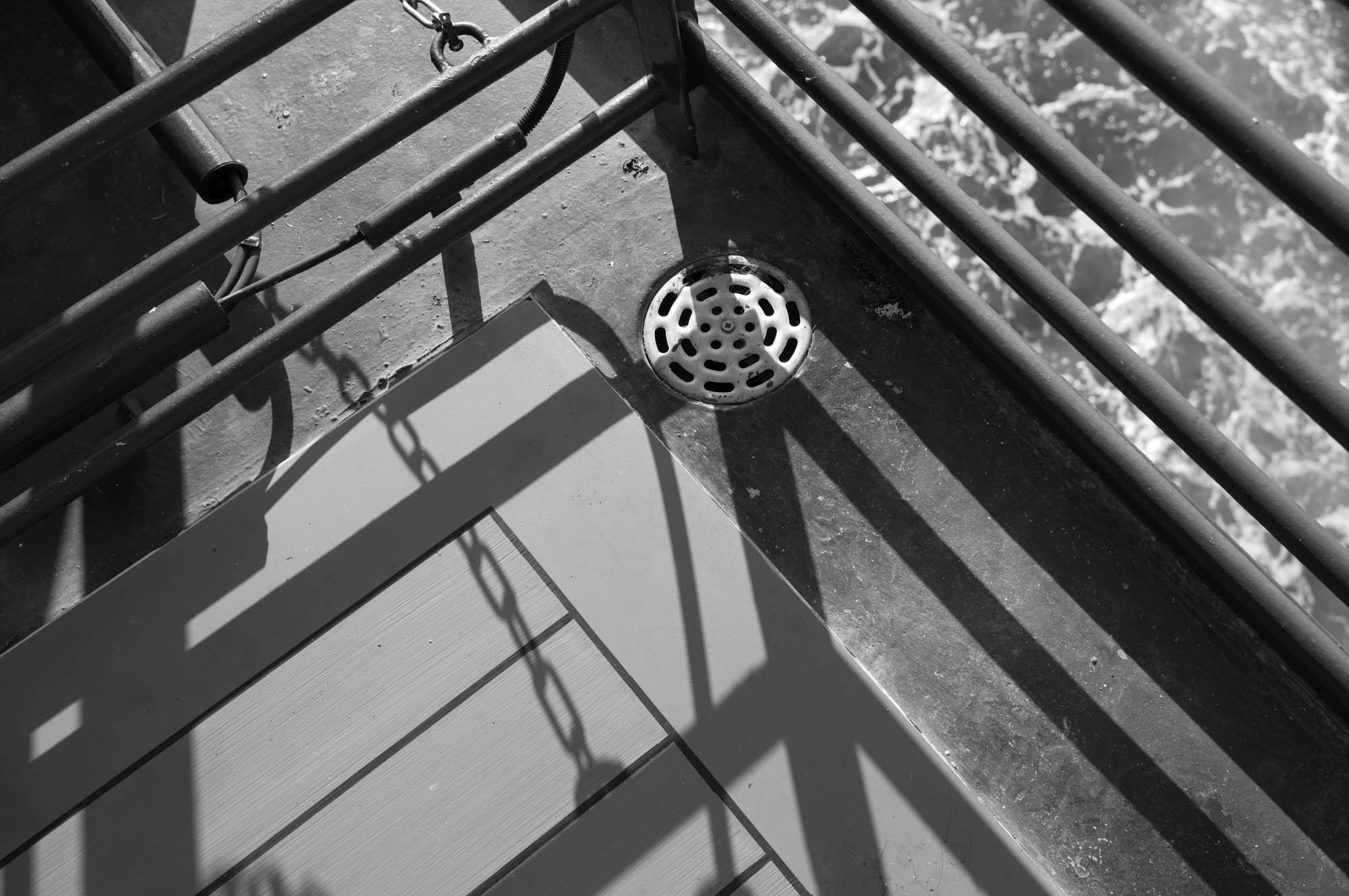 Black and white view of metal railings casting shadows near a drain on a ship's deck with visible water below.