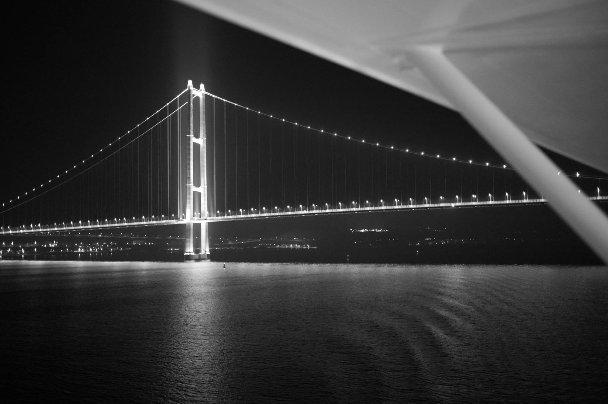 Illuminated suspension bridge at night across calm water, featuring glowing lights and city in the background. Black and white.