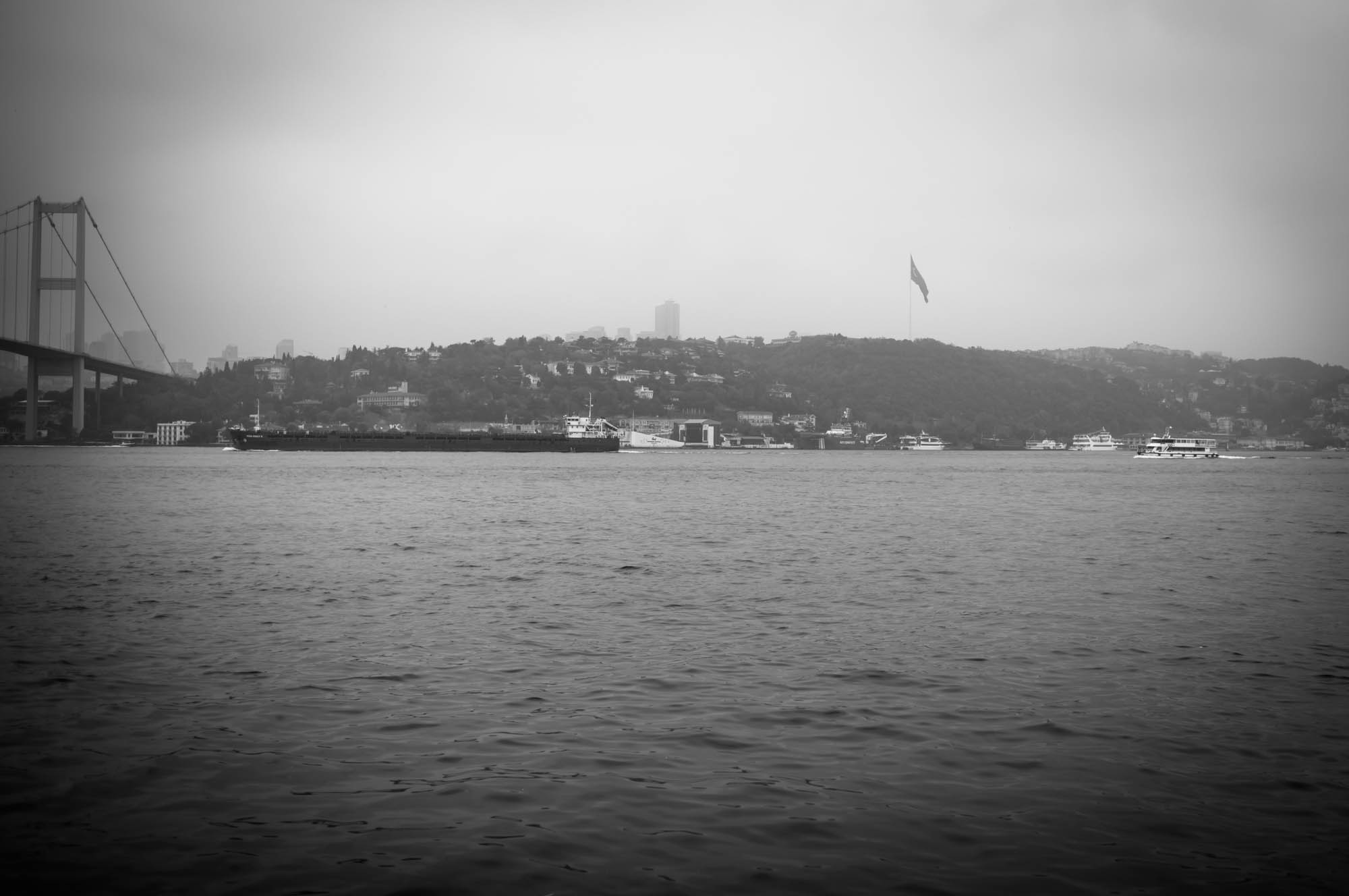 Monochrome view of a bridge over a river with ships, hilly landscape, and a large flag in the background.