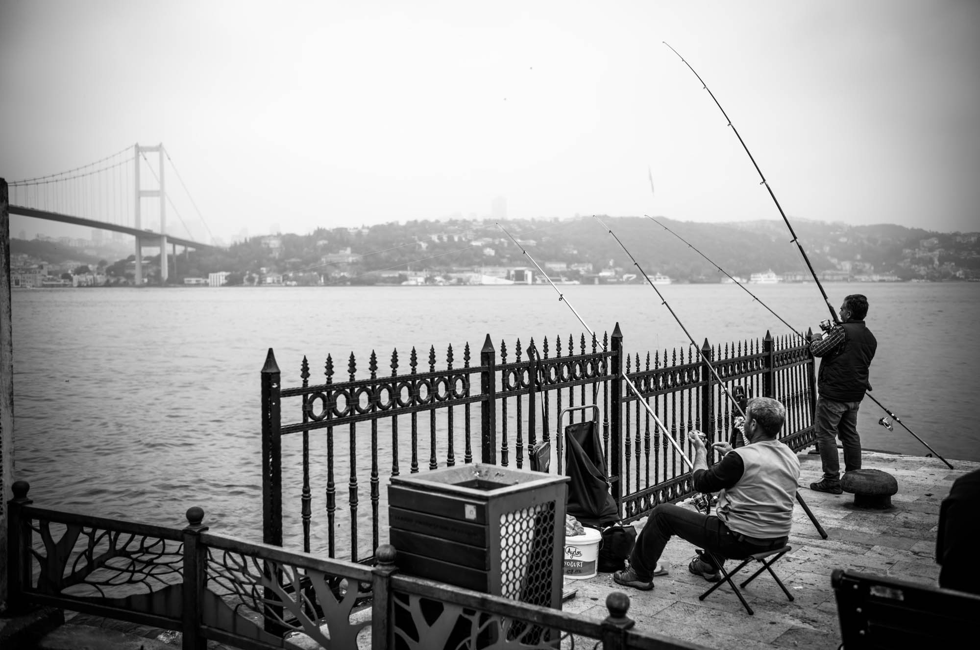 Two people fishing by a railing near a river, with a bridge and cityscape in the background.