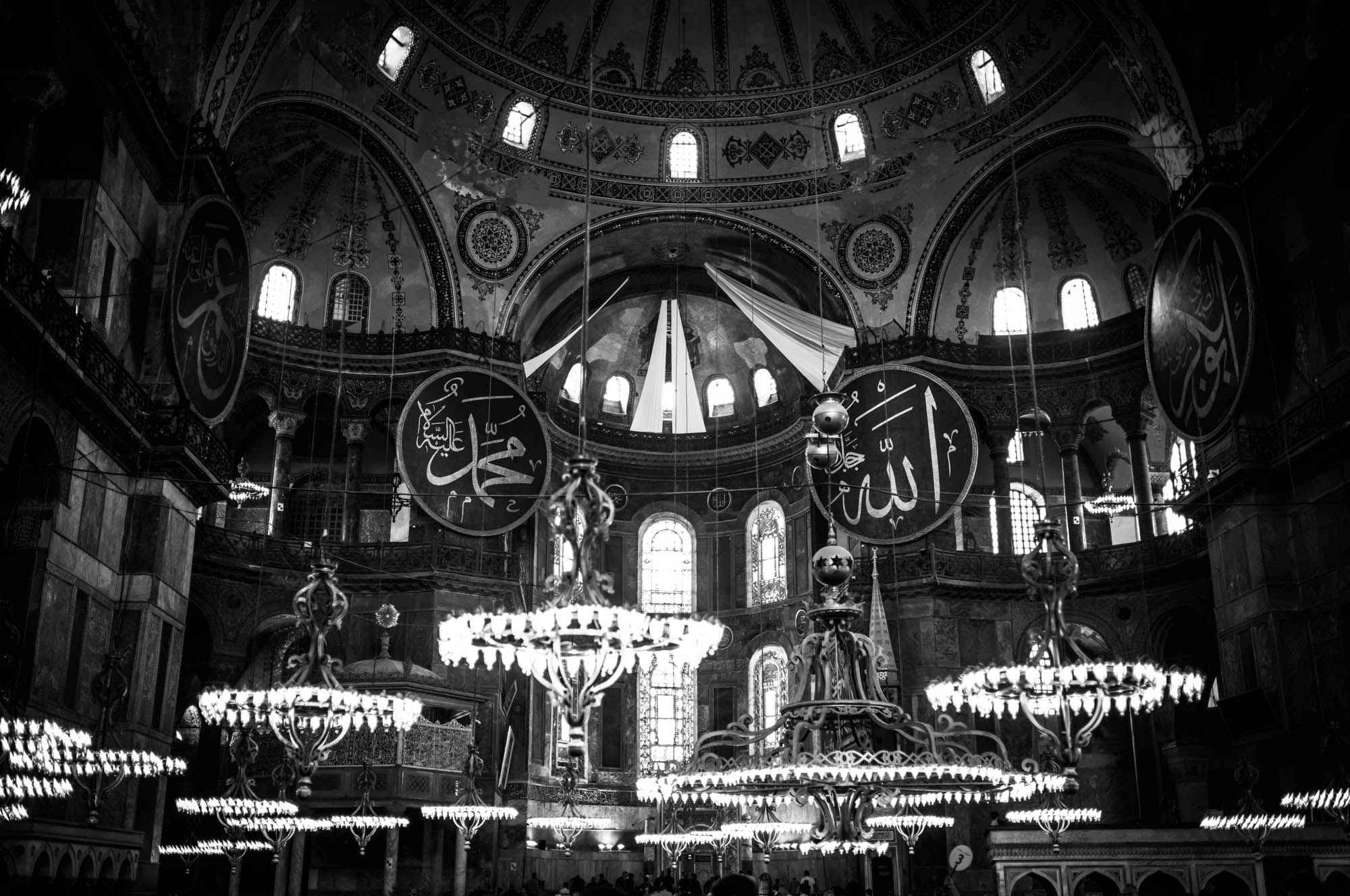 Intricate interior of Hagia Sophia with ornate chandeliers and Islamic calligraphy, captured in black and white.